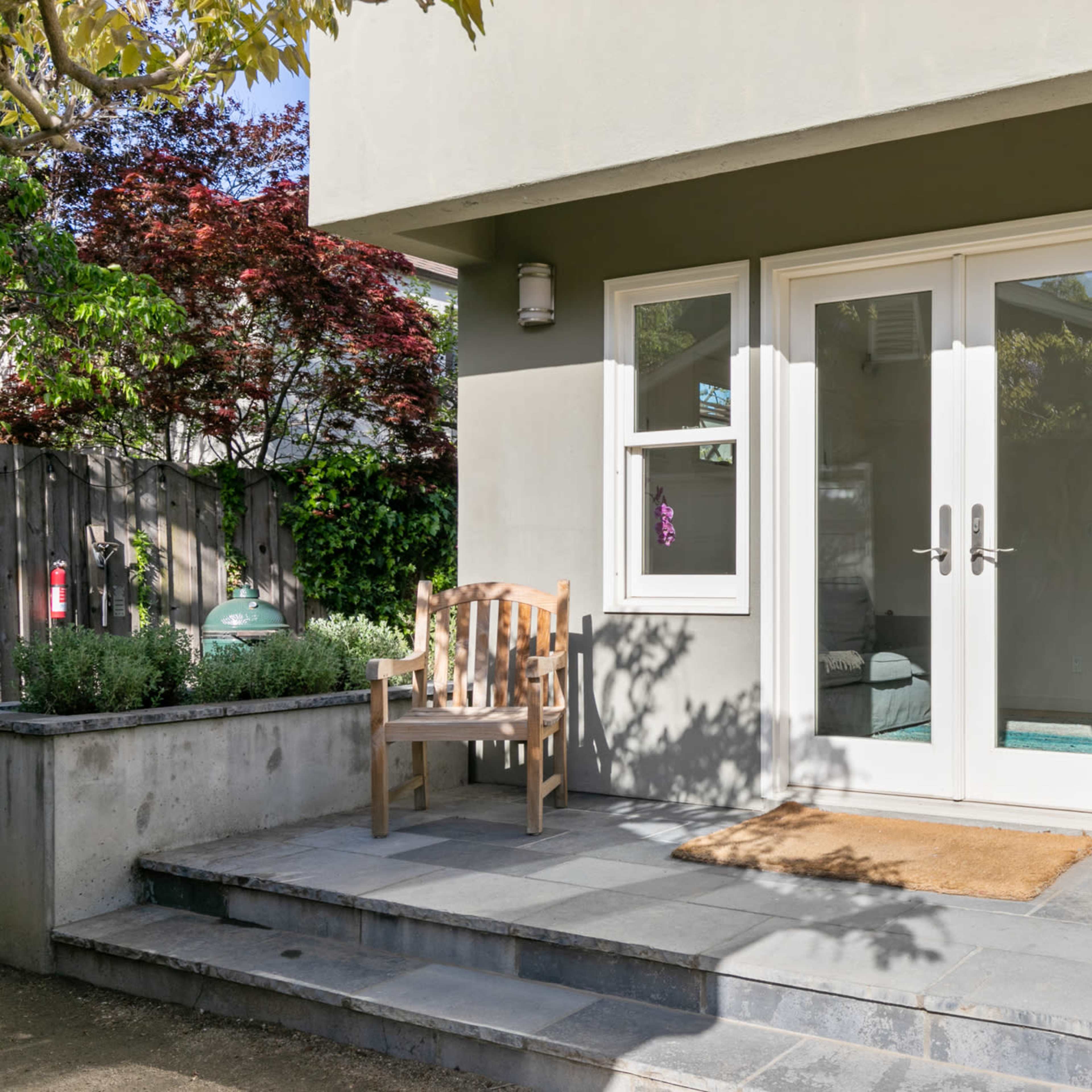 A minimalist entrance features a set of double doors, flanked by two windows, leading to a stone pathway and a wooden bench.