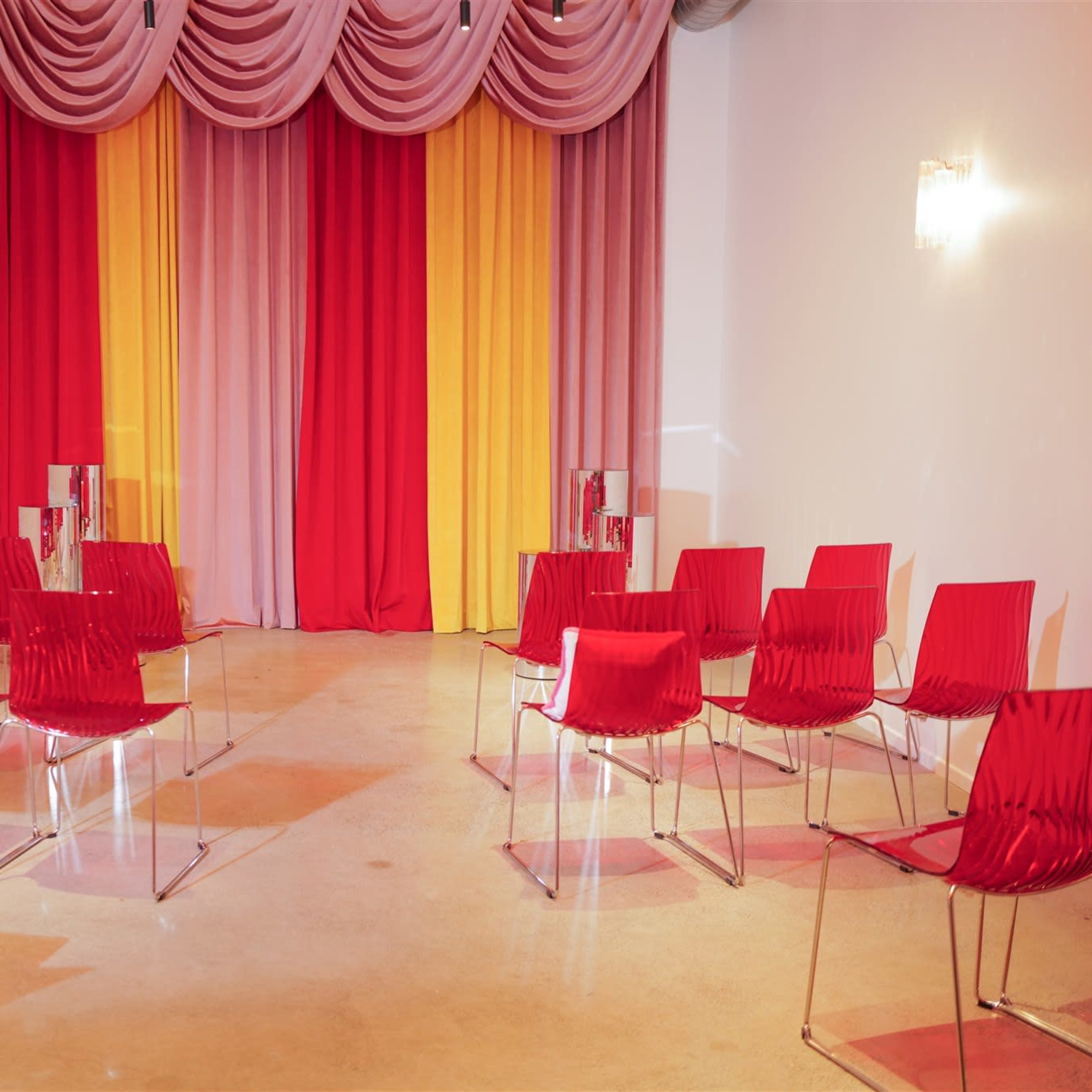 A room set up with several red chairs arranged in a semi-circle facing a backdrop of draped fabric in red, pink, and yellow.