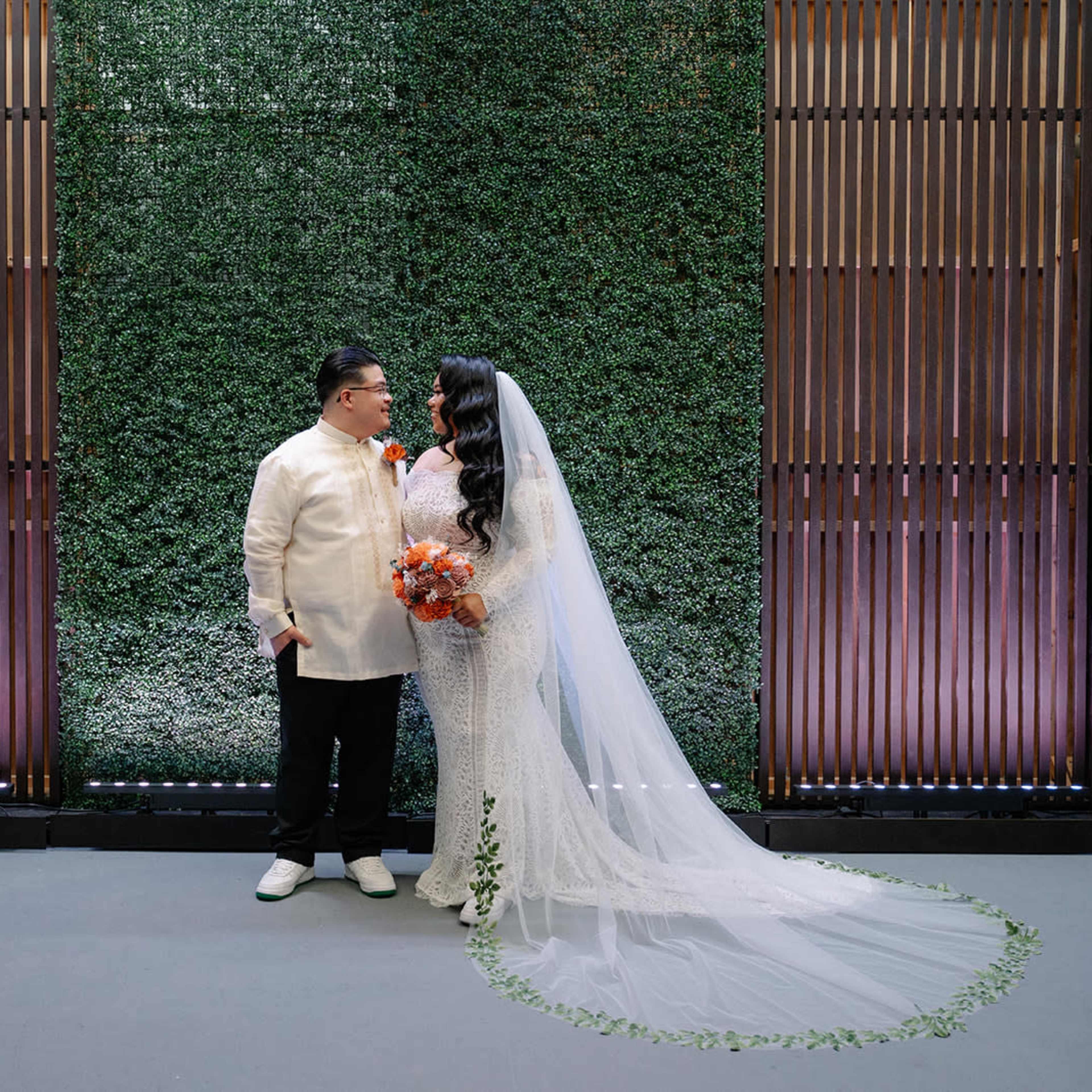 A bride in a white gown and veil stands next to a groom in traditional attire against a lush green backdrop.