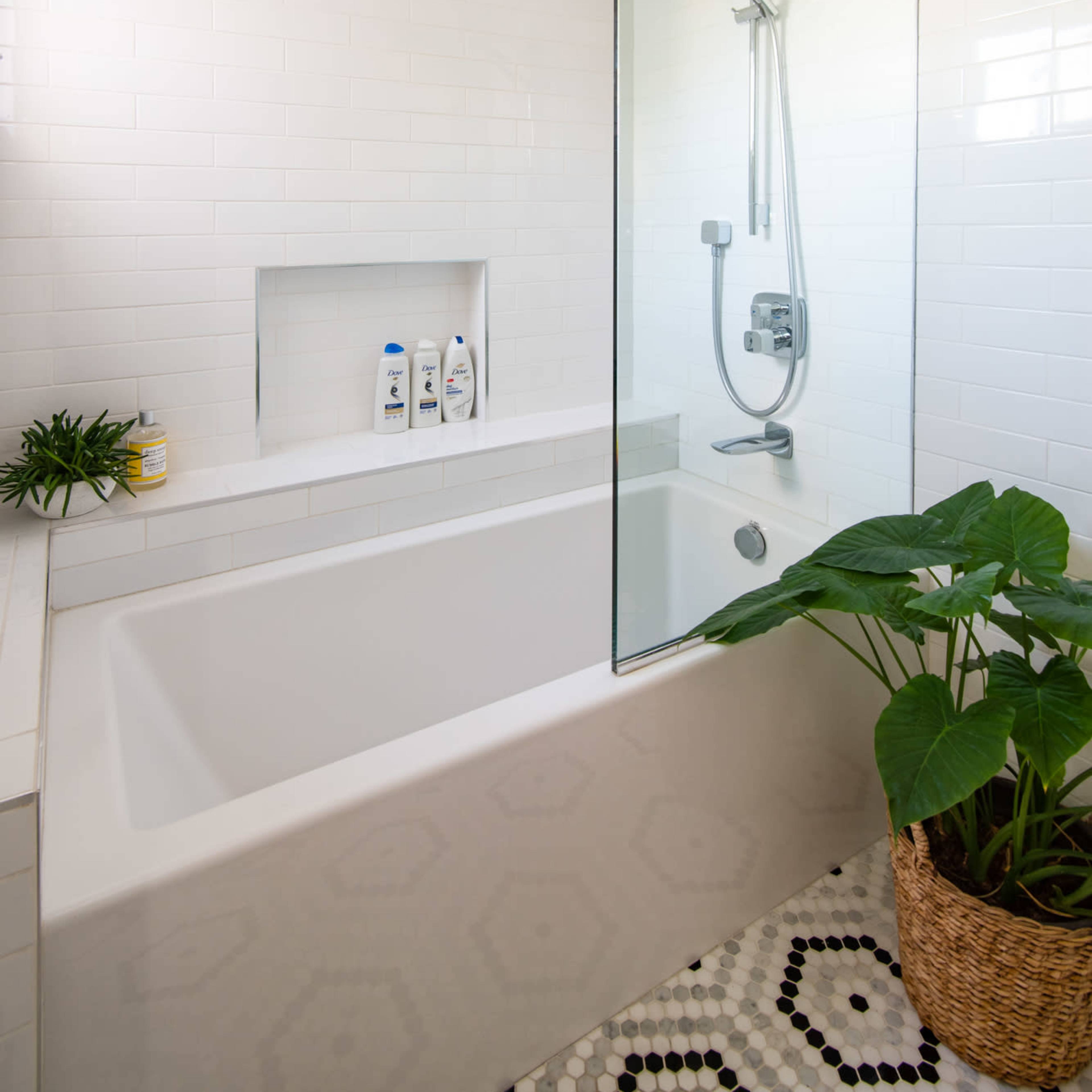 A modern bathroom featuring a bathtub with a glass shower partition, a built-in shelf for toiletries, and a potted plant in the corner.