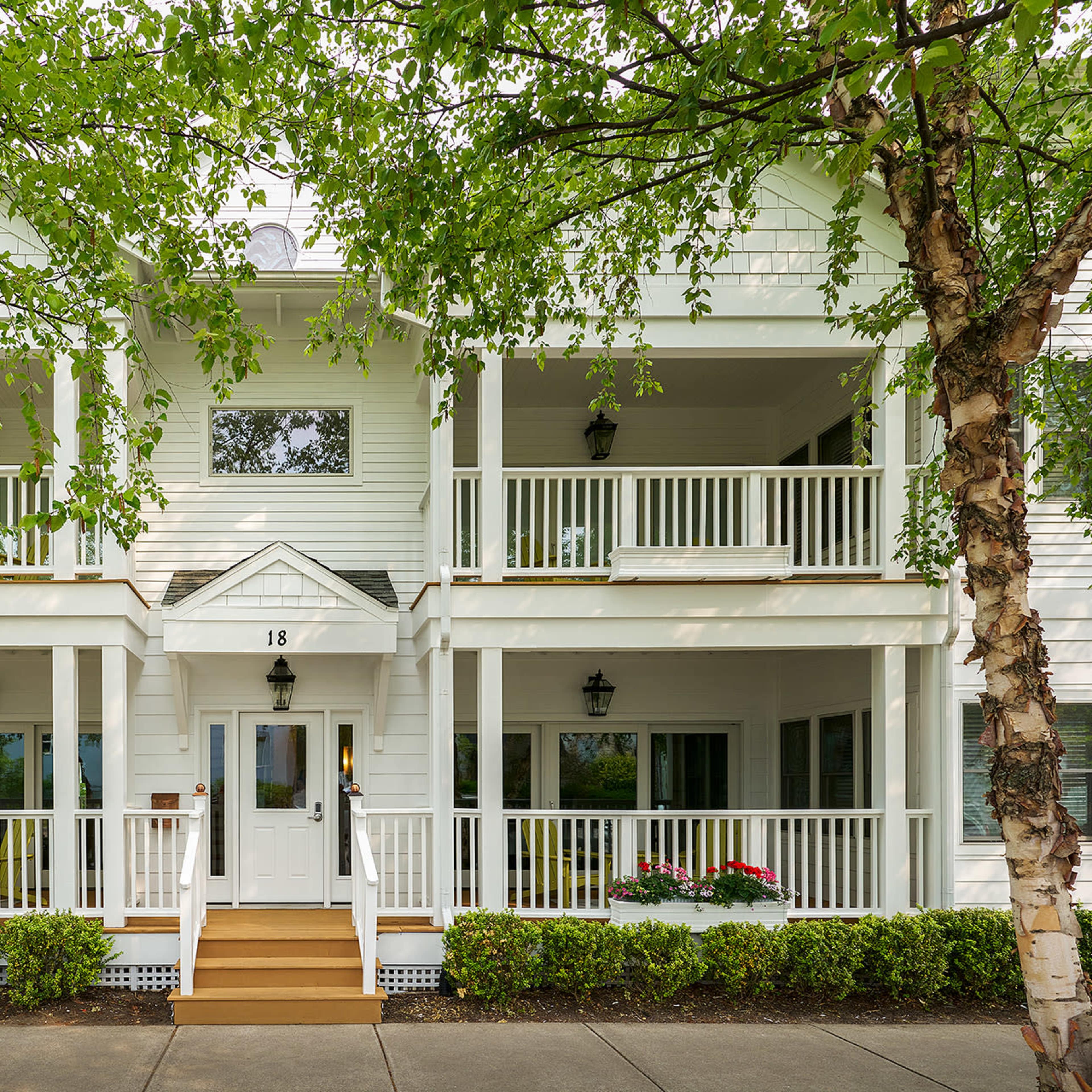 A white two-story house with multiple balconies, framed by trees, features a central entrance labeled "18" and flower beds on either side.