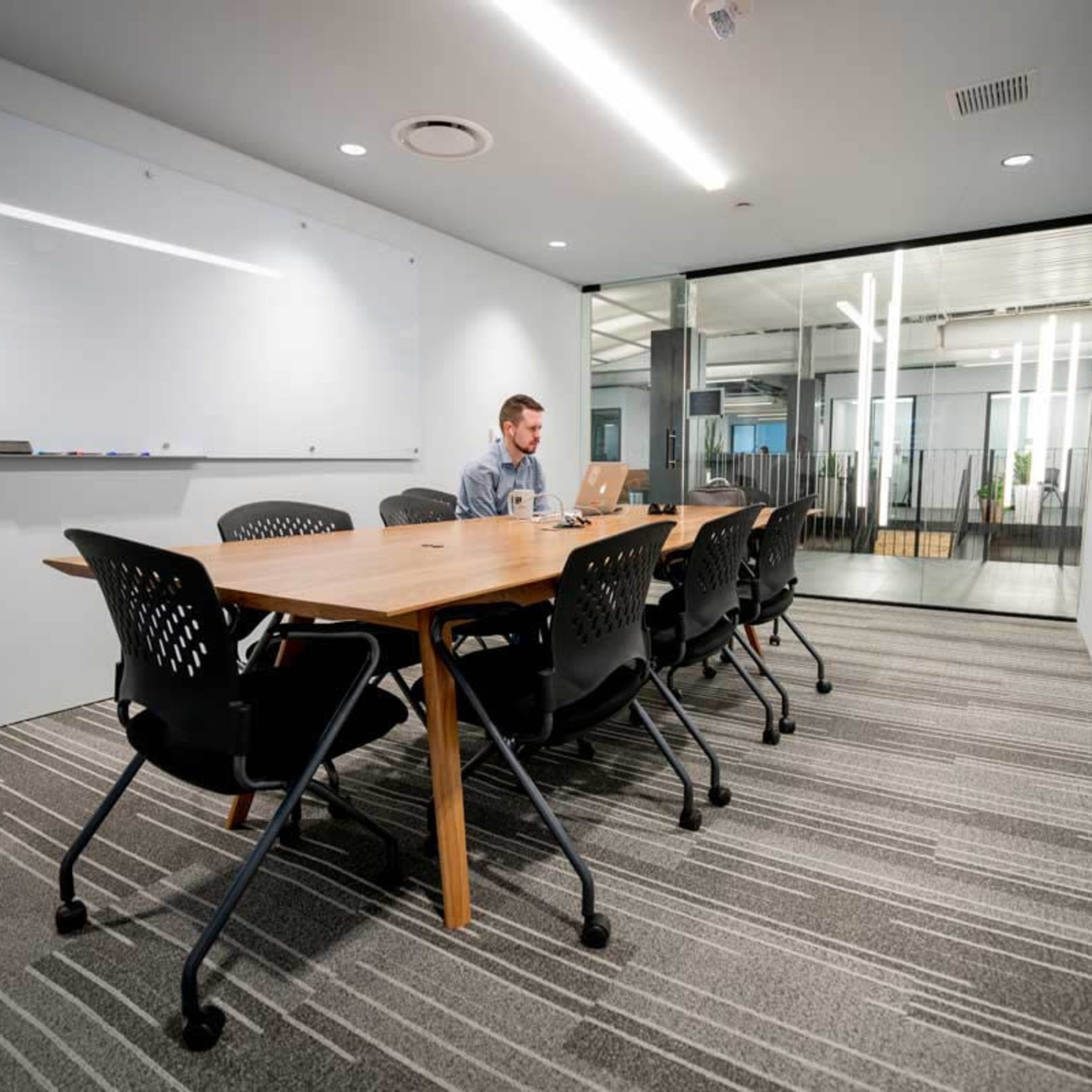 A man sits at a long wooden table in a modern conference room, working on a laptop while surrounded by empty chairs.