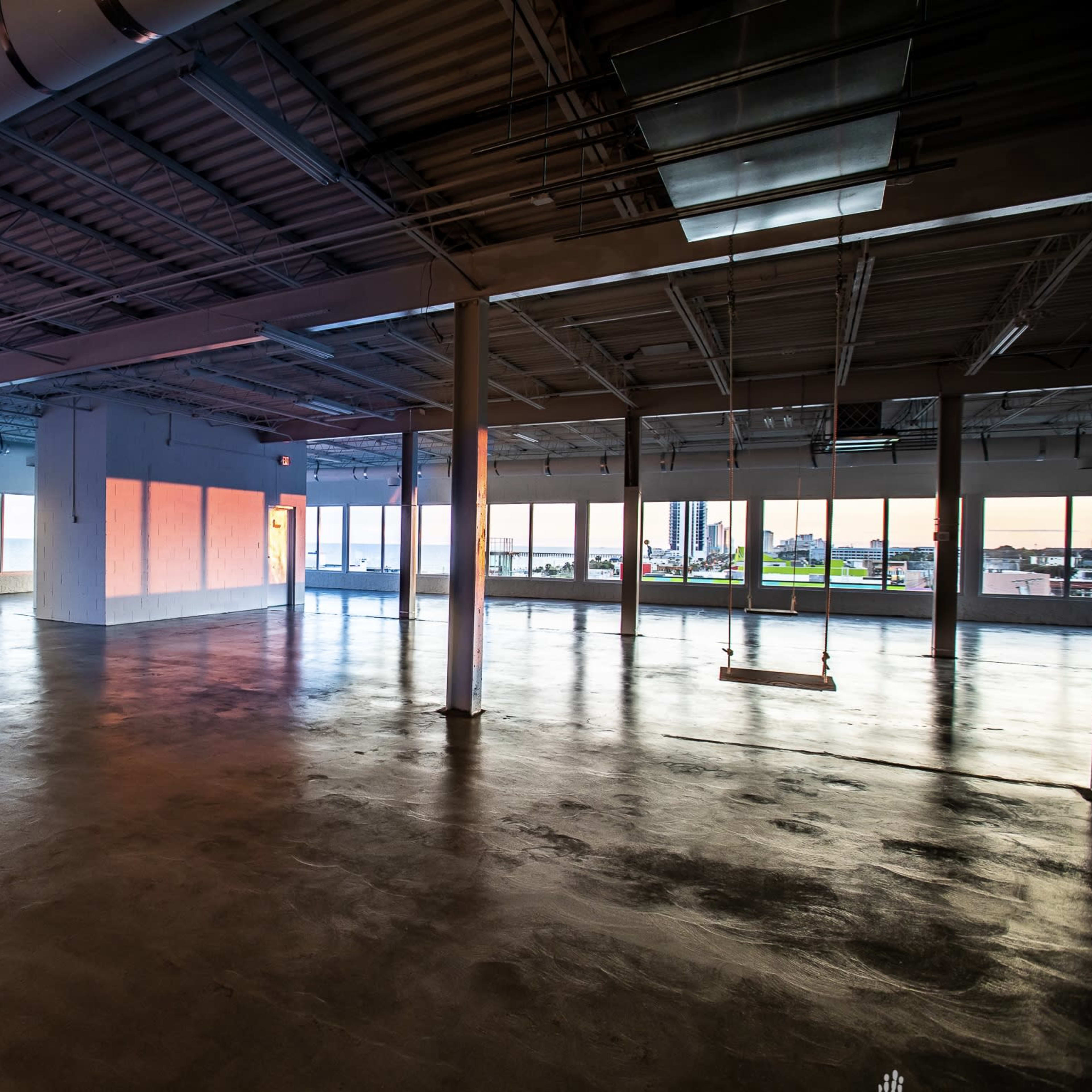 A spacious, empty room with large windows overlooking a body of water and a cityscape, illuminated by soft evening light.