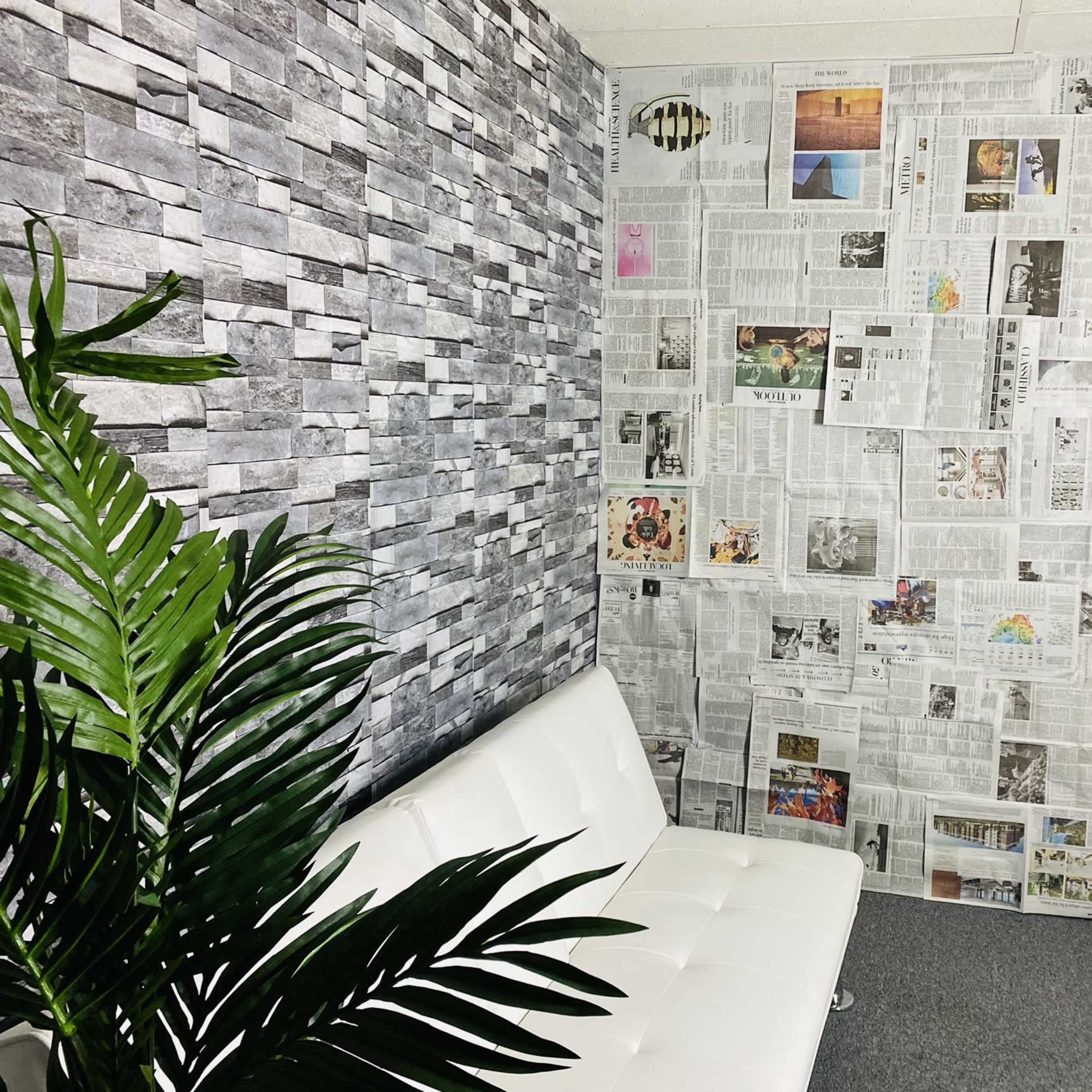 A room with one wall covered in gray stone wallpaper and the opposite wall decorated with newspaper clippings, featuring a white sofa and a green plant in the corner.