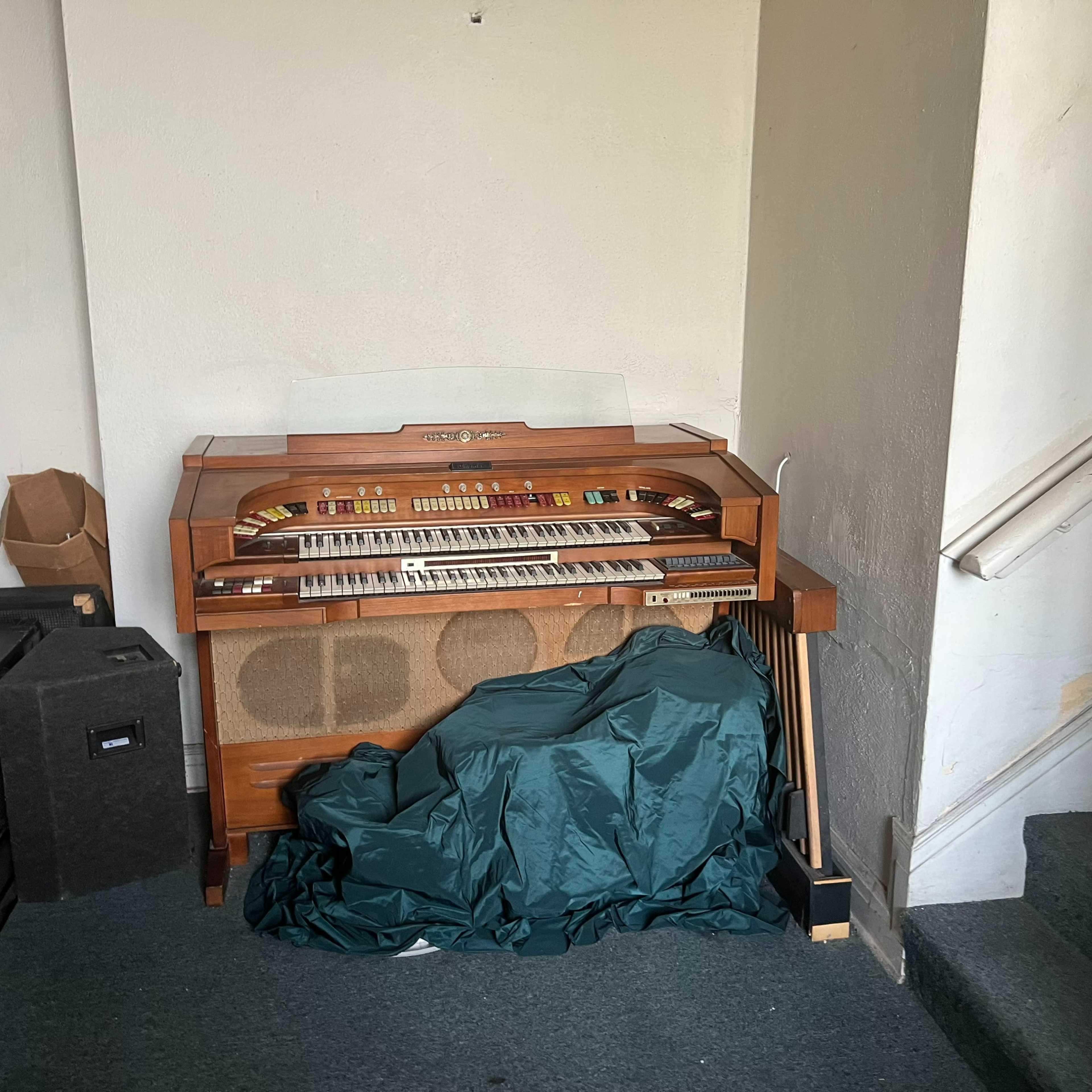 A large organ is partially concealed by a green cover in a corner of a room beside various musical equipment and a staircase.