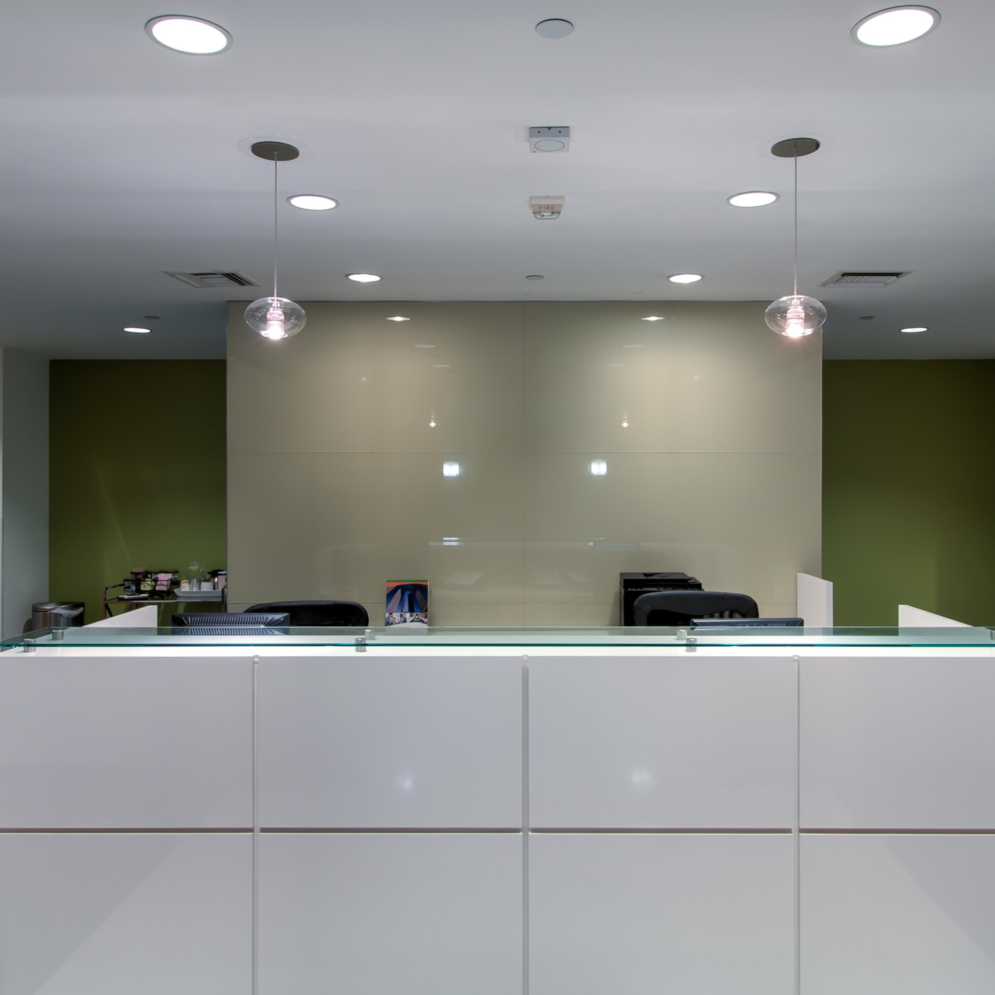 A modern reception area features a white desk with a glass top, backed by a green wall and simple overhead lighting.