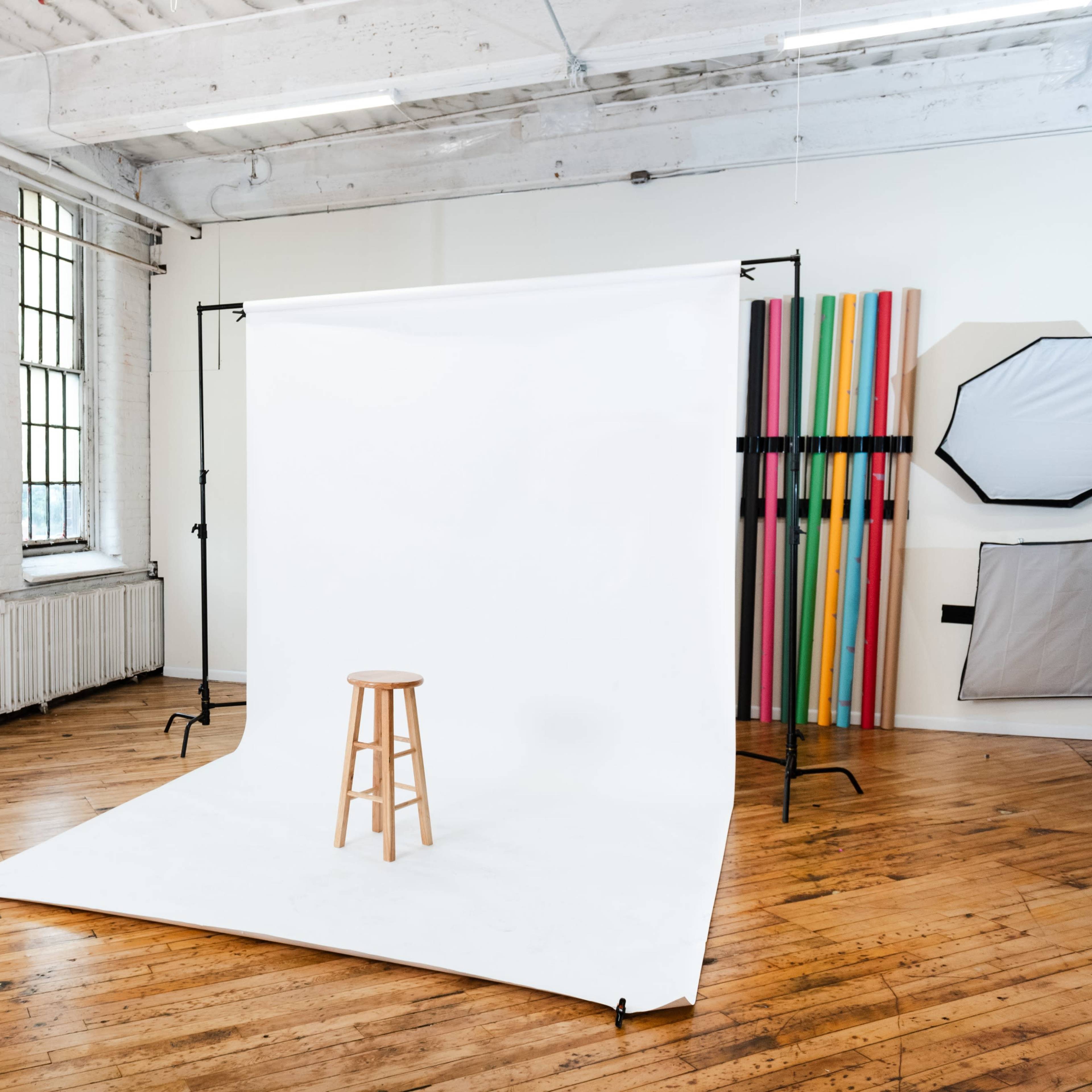 A photography studio with a wooden stool positioned on a white backdrop, surrounded by large windows and colorful props.