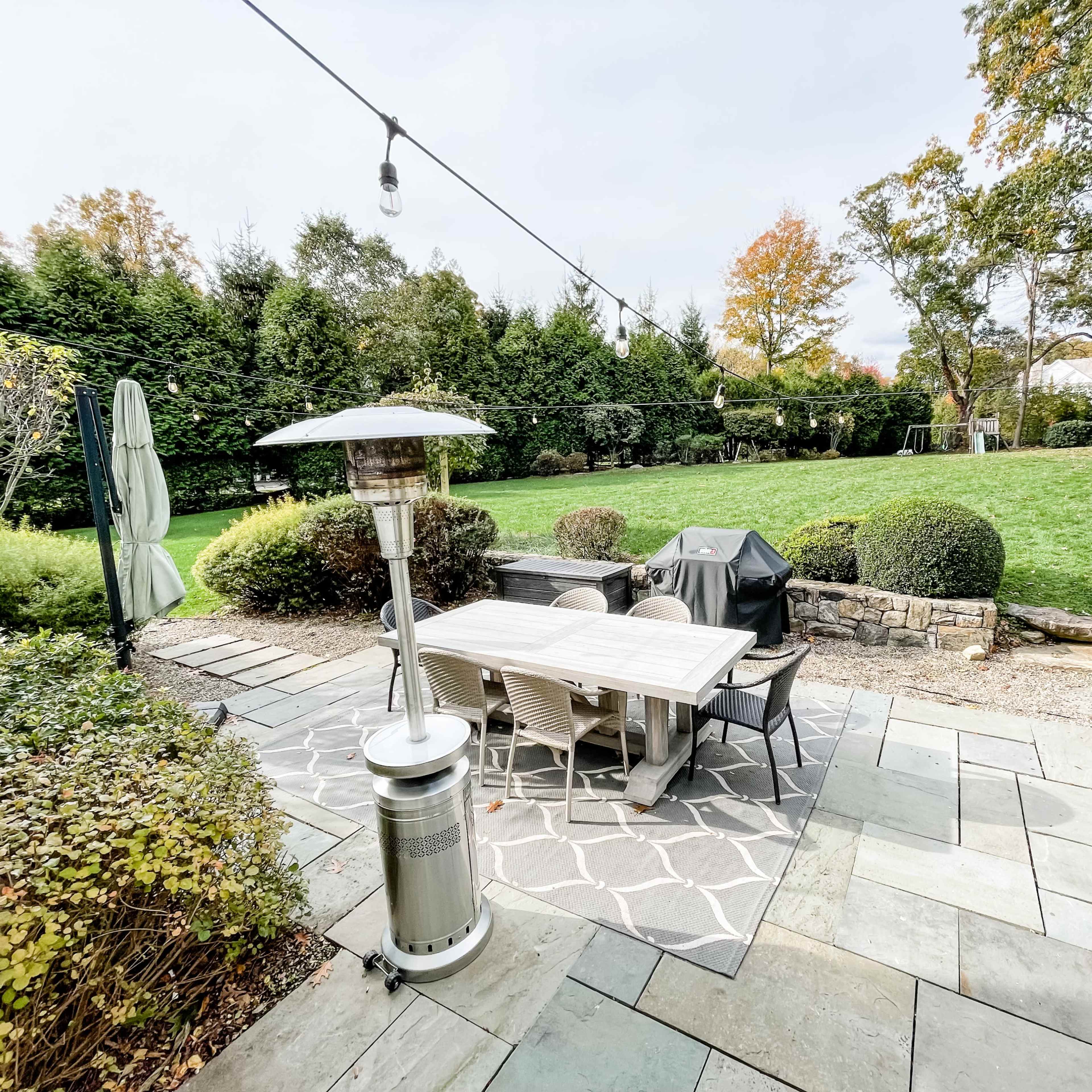 A stone patio with a table and chairs under a patio heater, surrounded by neatly trimmed hedges and a large green lawn.
