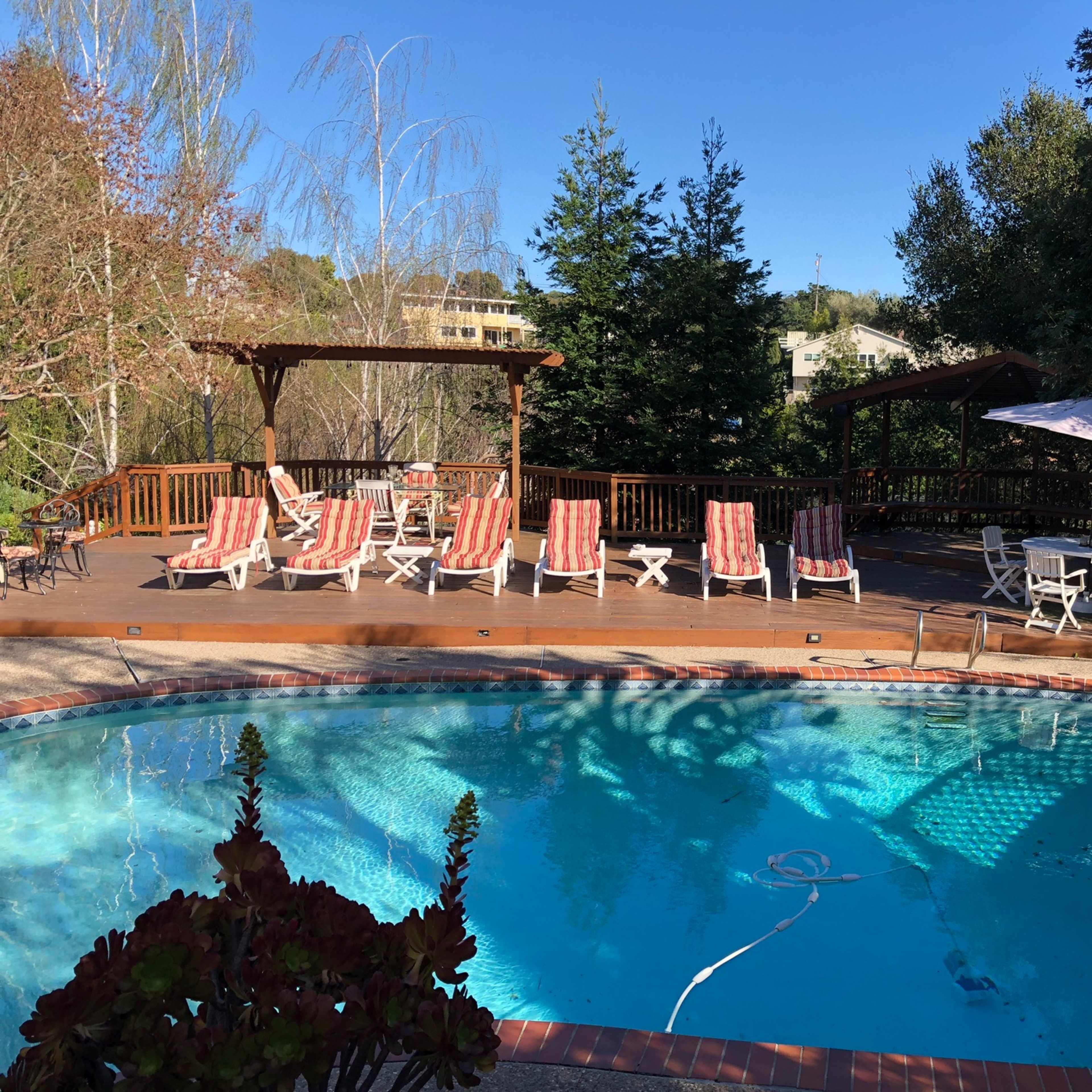 A swimming pool with a wooden deck surrounded by lounge chairs and tables under shaded structures.