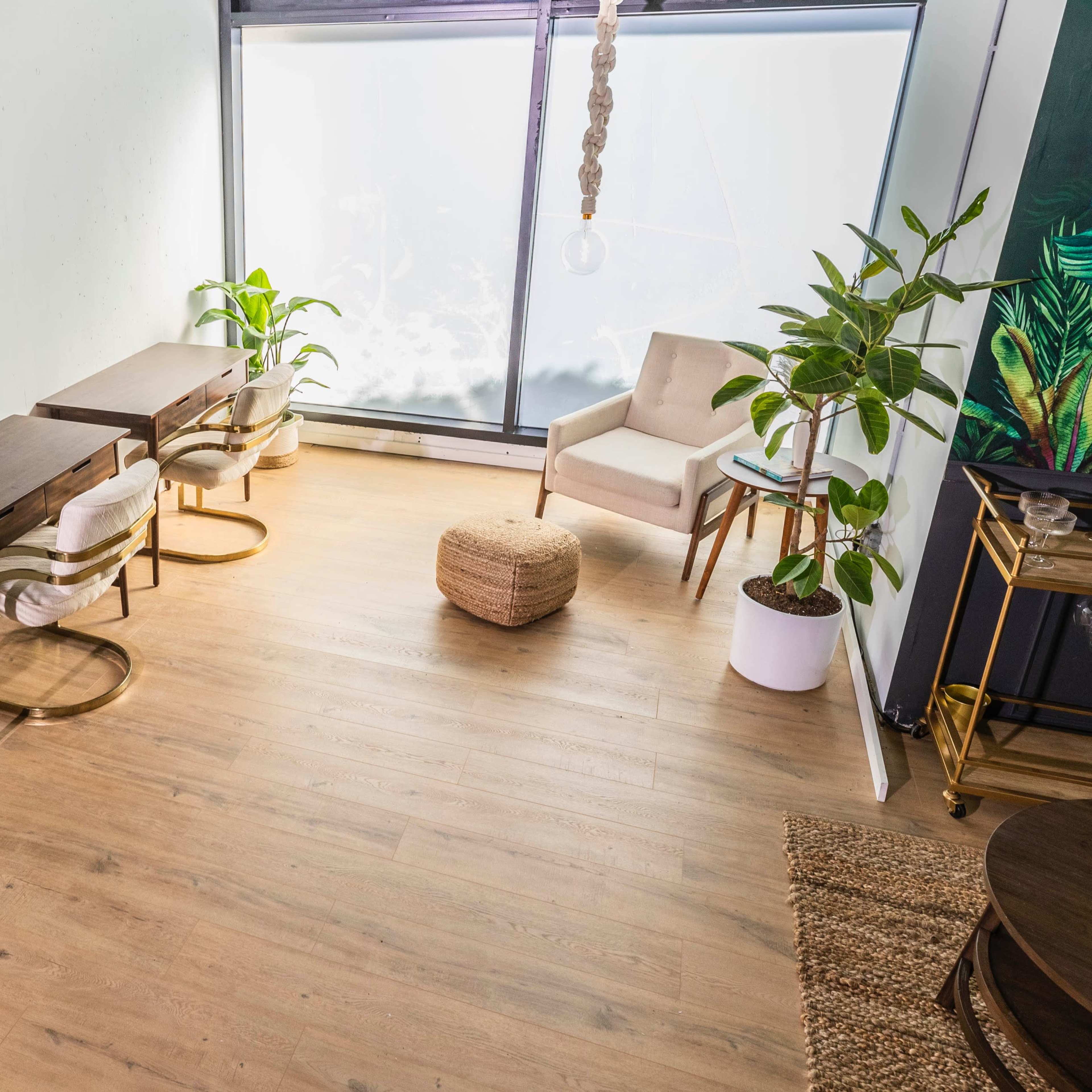 A bright, minimalistic room with two wooden tables, several indoor plants, a beige chair, and a textured pouf on a wooden floor near a large window.