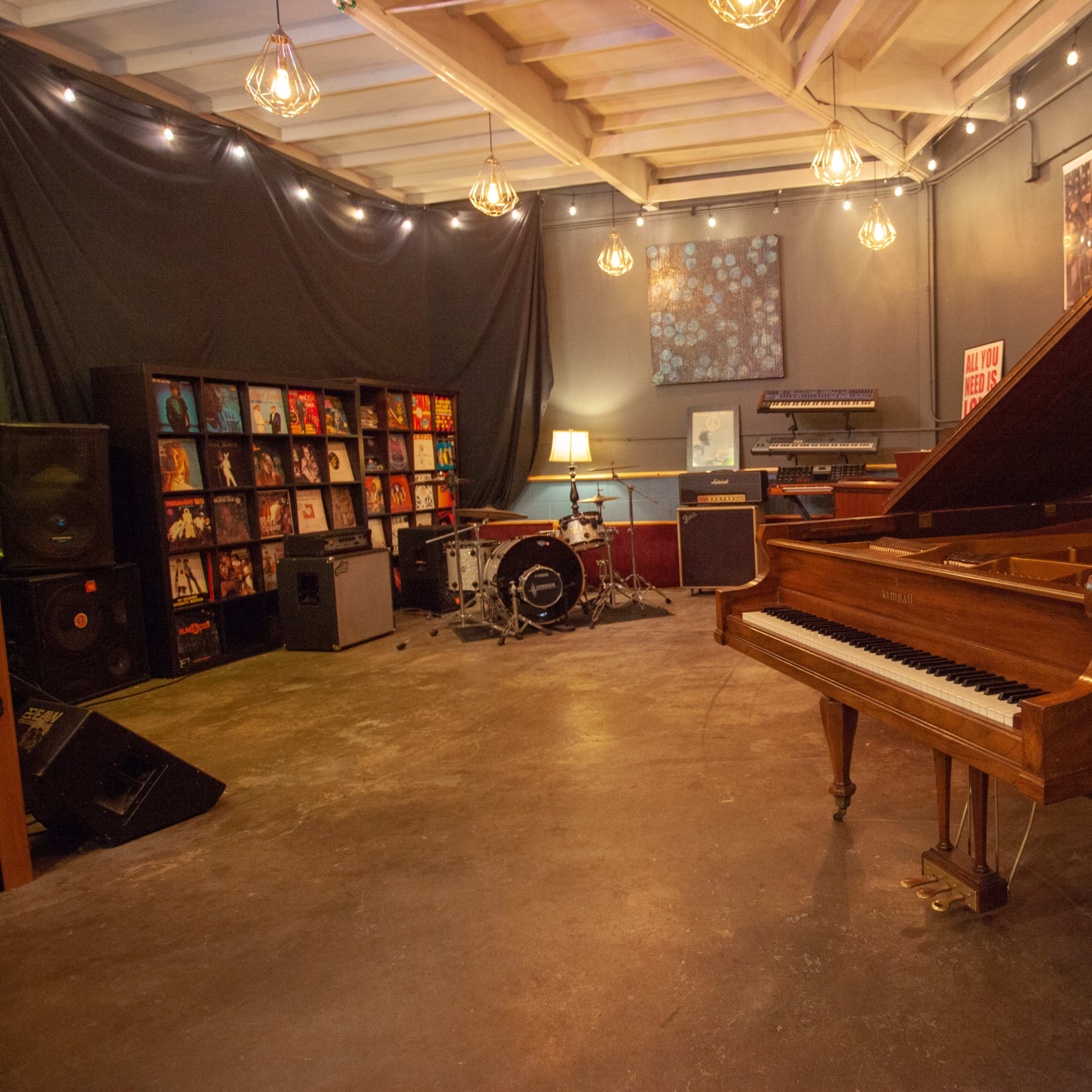 A music studio featuring a grand piano, a drum set, speakers, record shelves, and warm overhead lights.