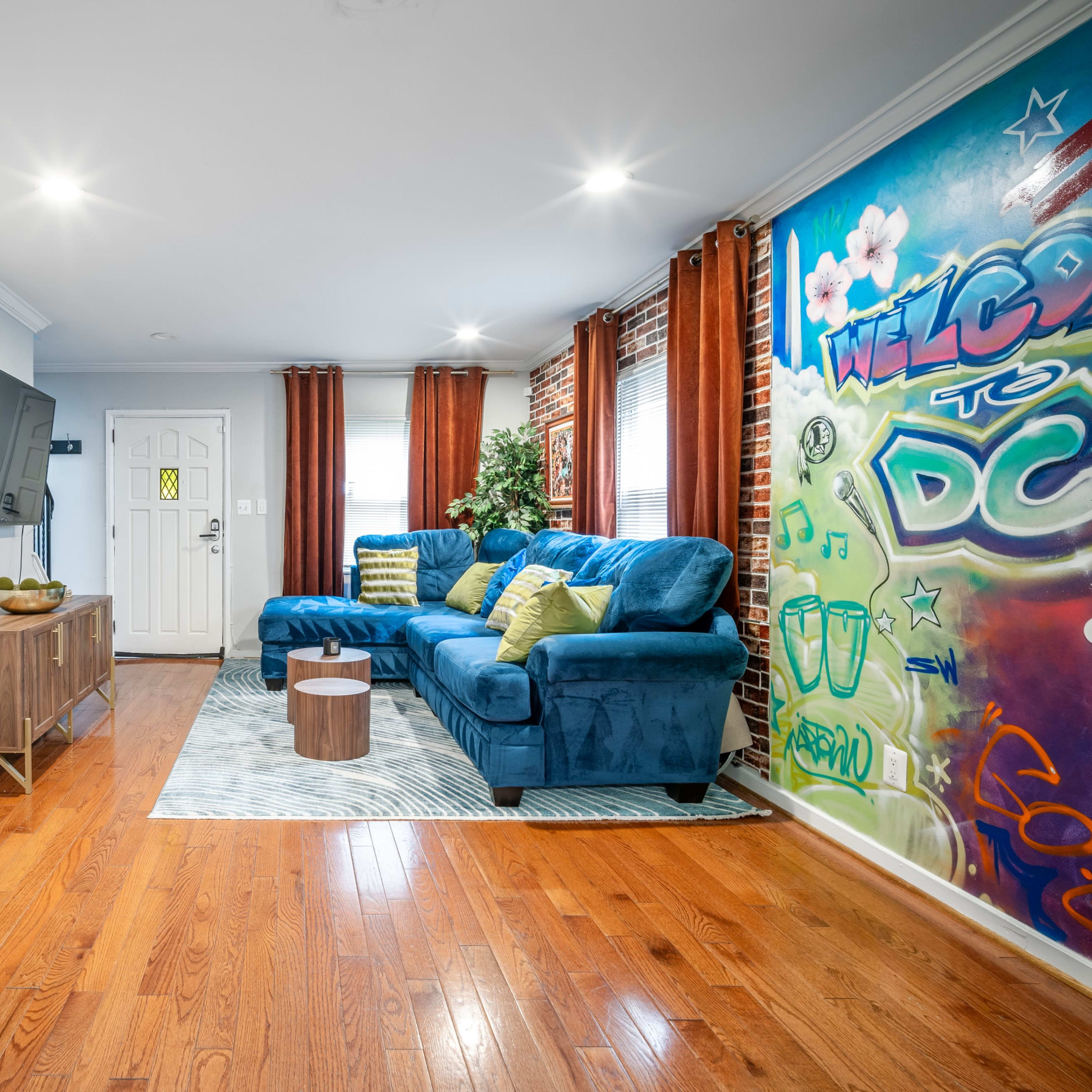 A modern living room featuring a blue sectional sofa, a wooden TV stand, and a colorful mural on a brick wall.