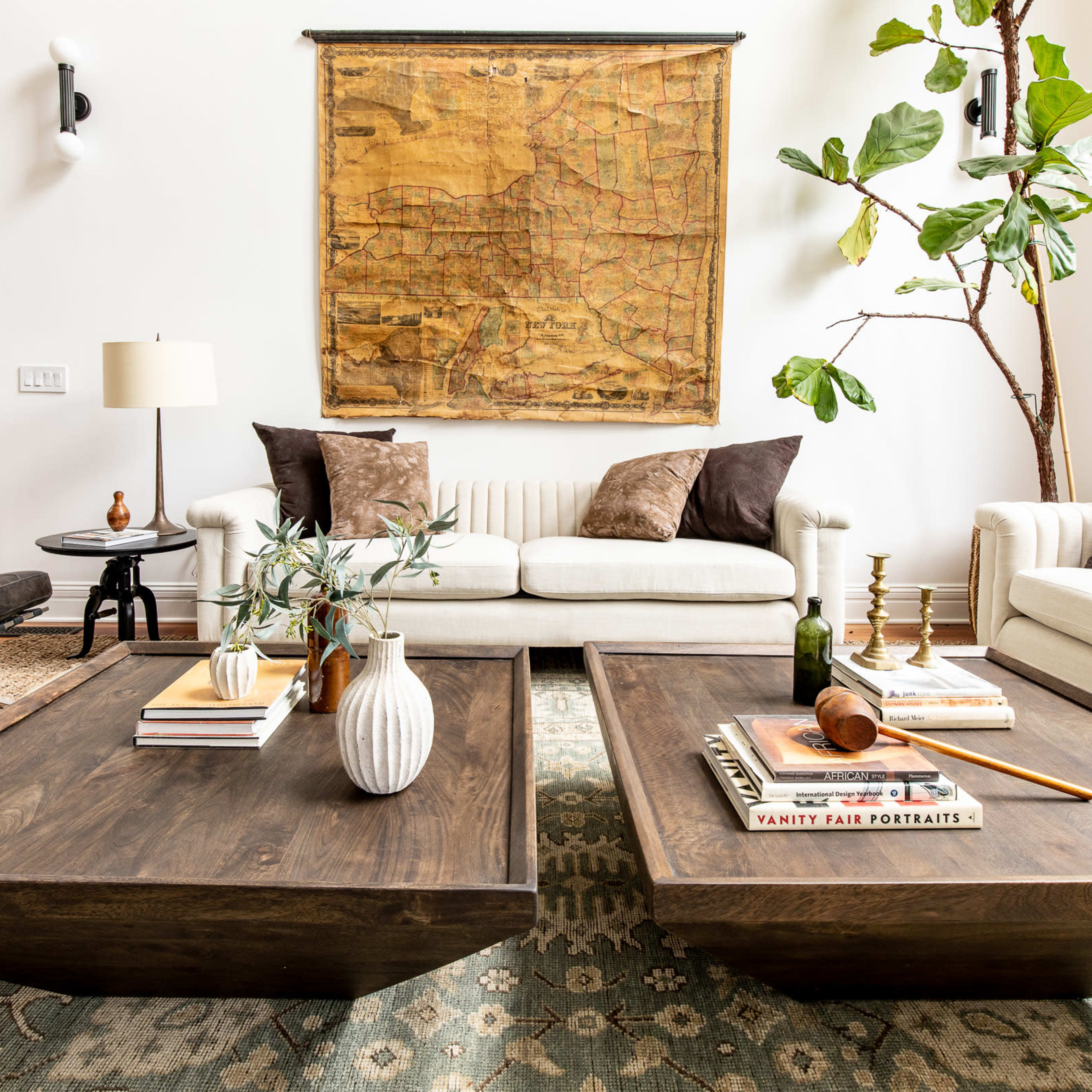 A living room features two low coffee tables with books and decor, surrounded by a sofa and a leafy plant, against a wall adorned with a large vintage map.