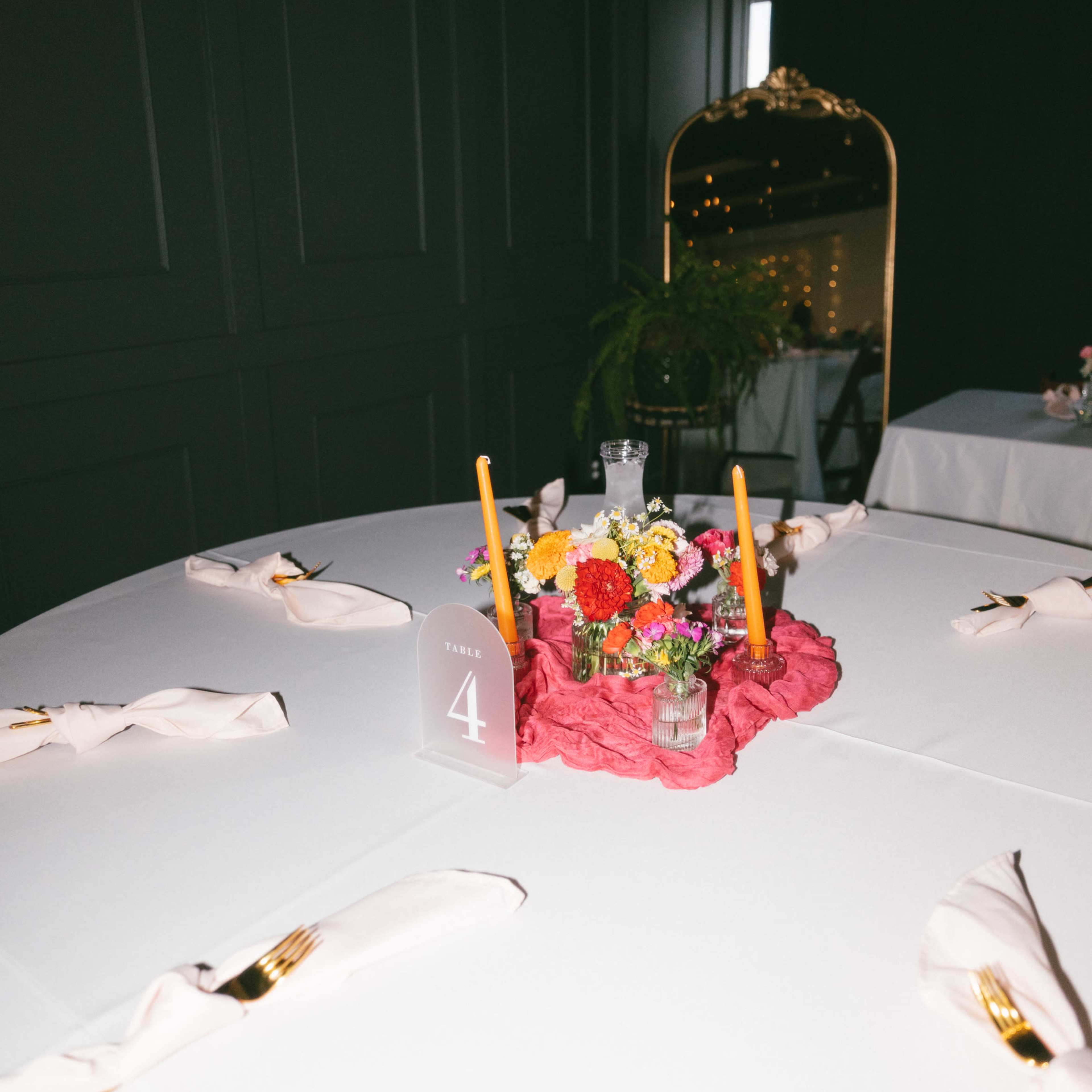 A round table is set with white linens and a centerpiece of colorful flowers and candles, surrounded by neatly arranged napkins and cutlery.