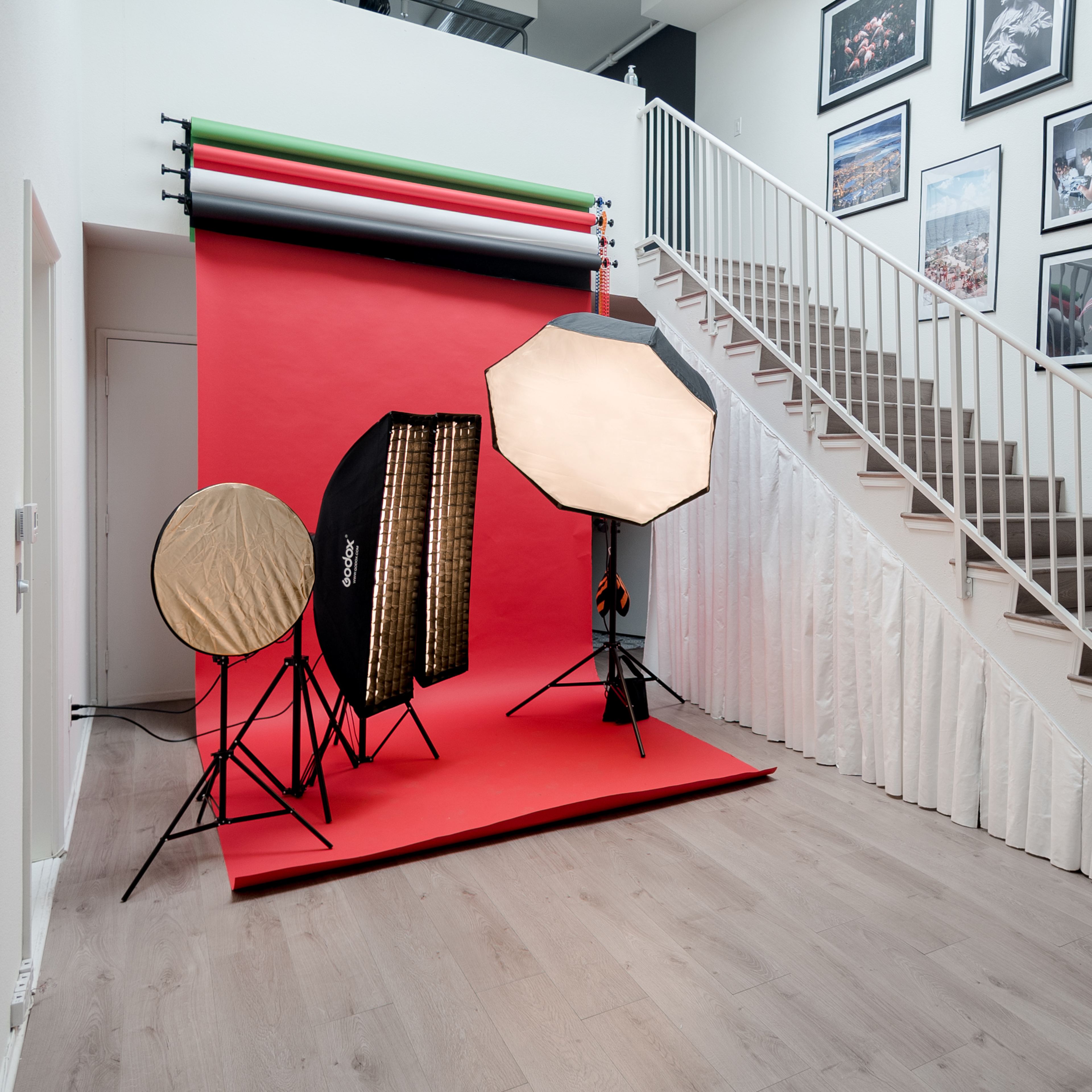 A photography studio setup with lighting equipment positioned against a red backdrop, located next to a staircase lined with framed photographs on the wall.