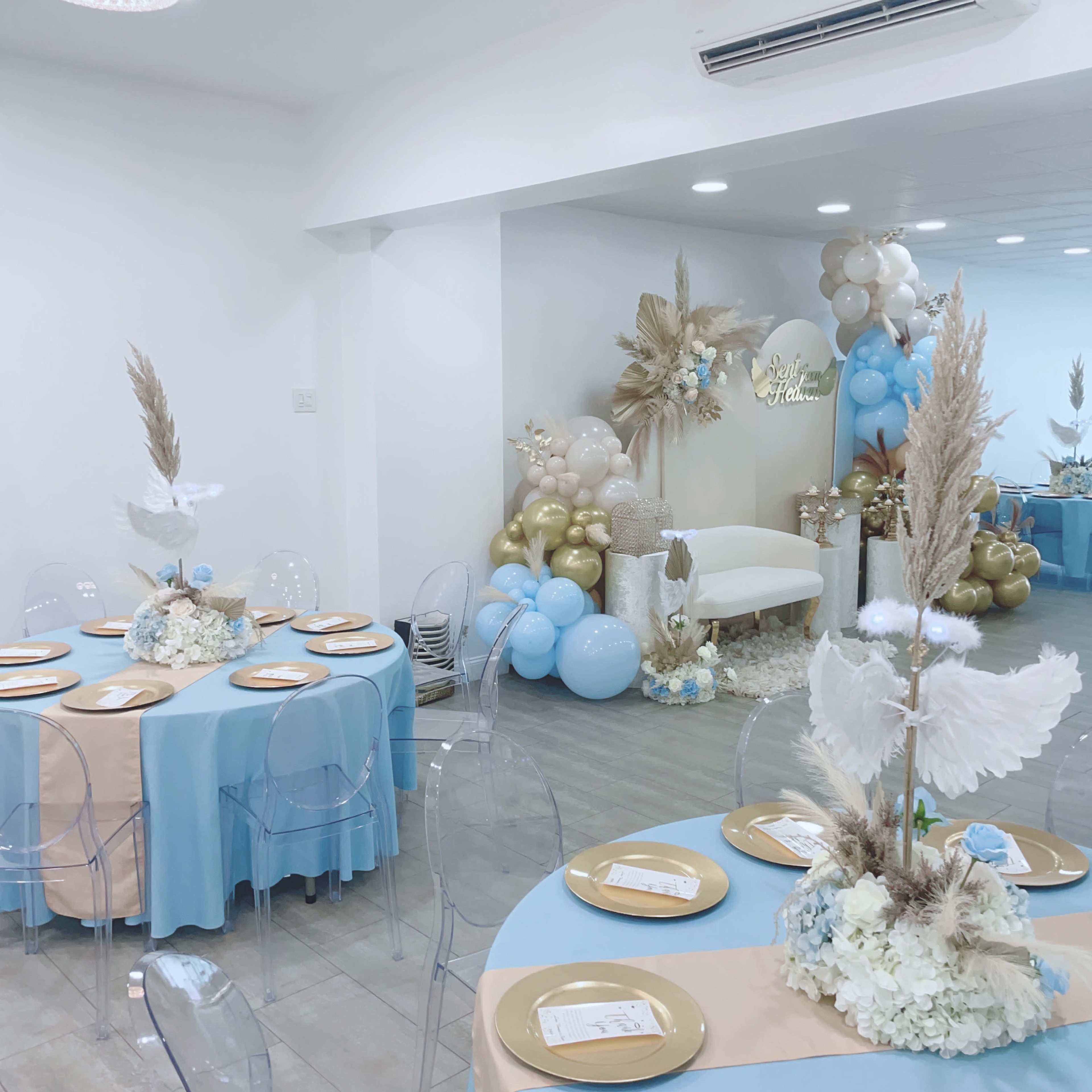 A decorated event space featuring round tables with blue and gold table settings, clear chairs, and a backdrop of balloons and dried flowers.