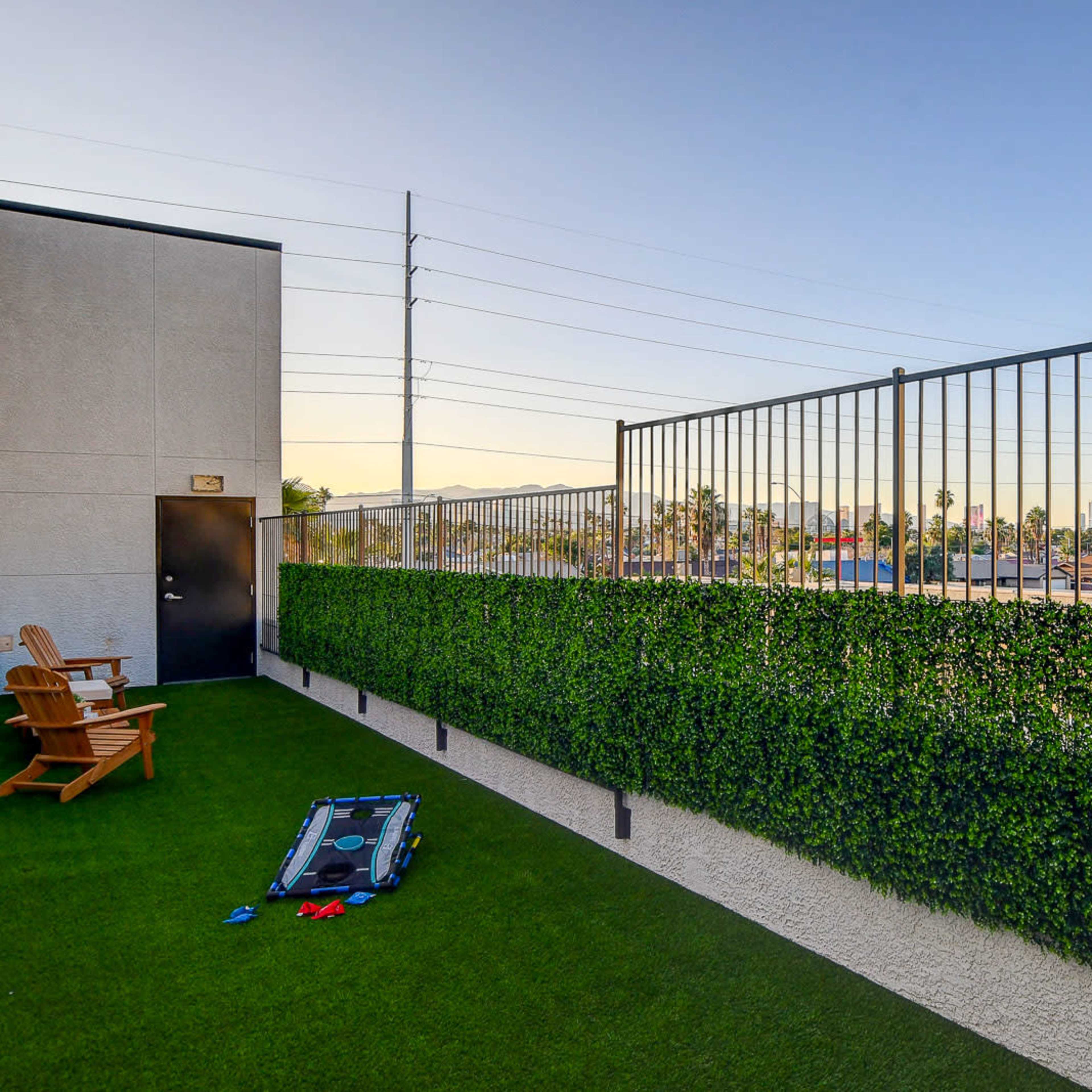 A rooftop balcony with artificial grass, a green privacy wall, and a view of distant buildings and power lines.