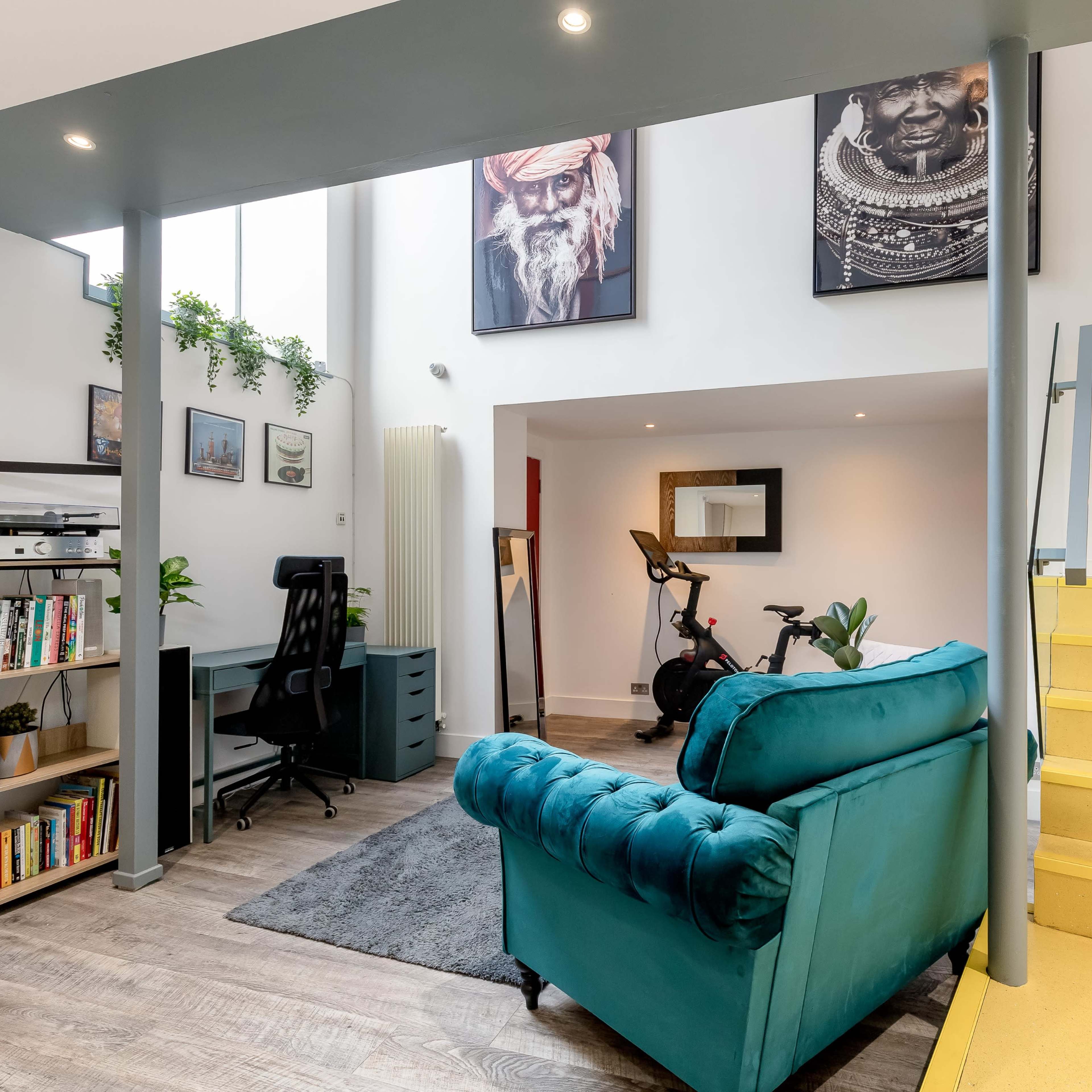 A modern interior featuring a turquoise couch, a desk with a chair, bookshelves, a yellow staircase, and wall art.