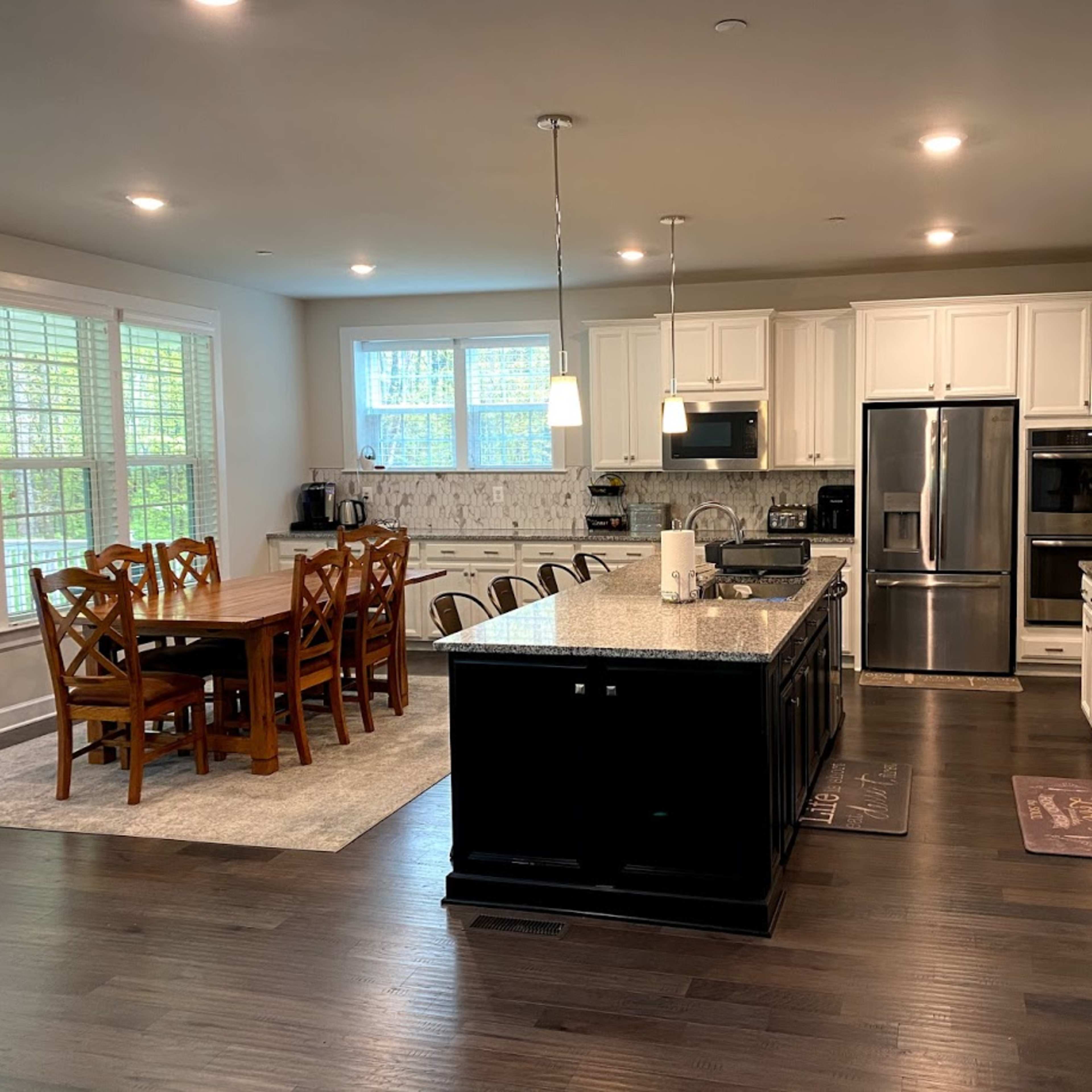 A modern kitchen with white cabinets, a large island with dark granite countertops, a dining table with six wooden chairs, and stainless steel appliances.