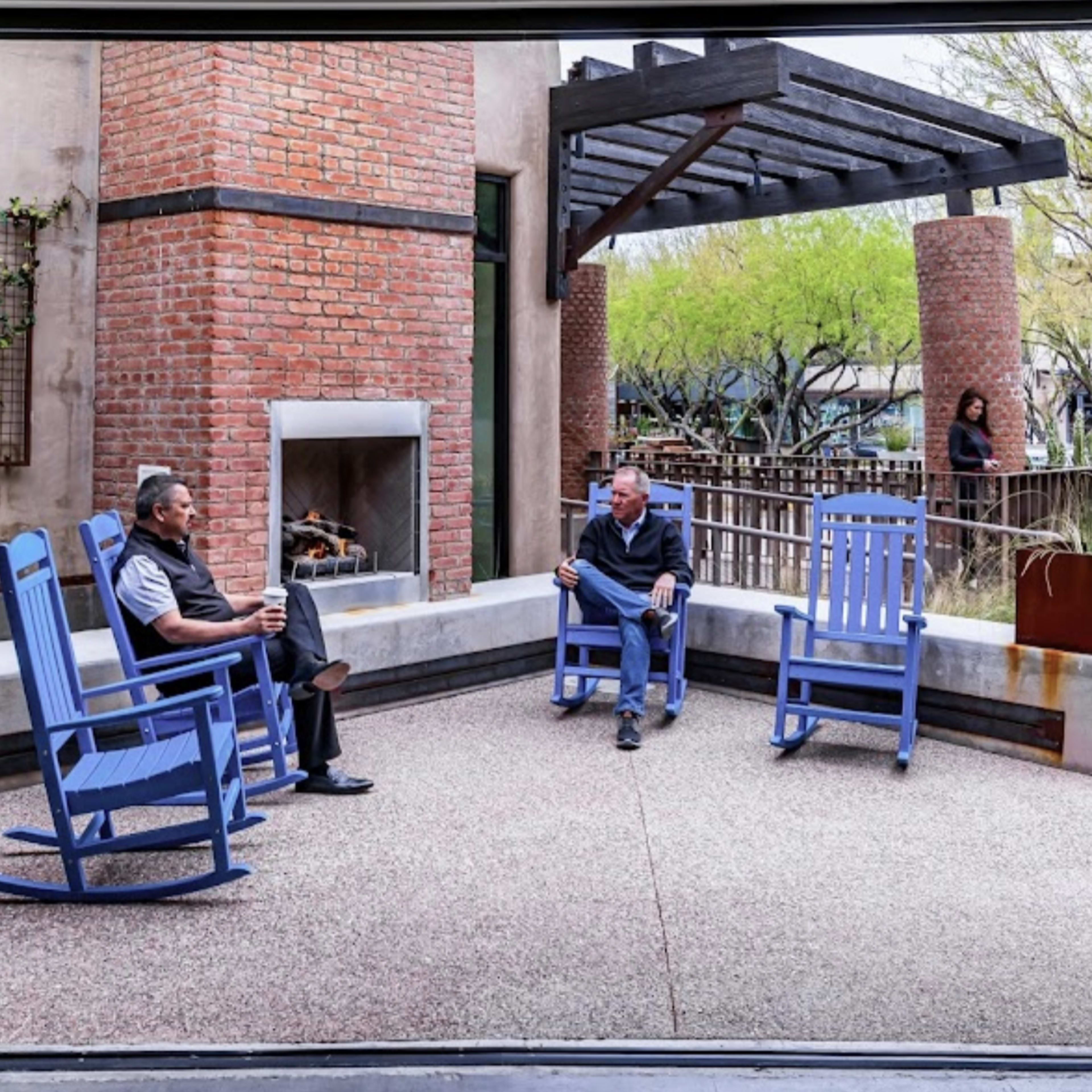 Two men sit in blue rocking chairs on a patio next to a brick fireplace surrounded by greenery.