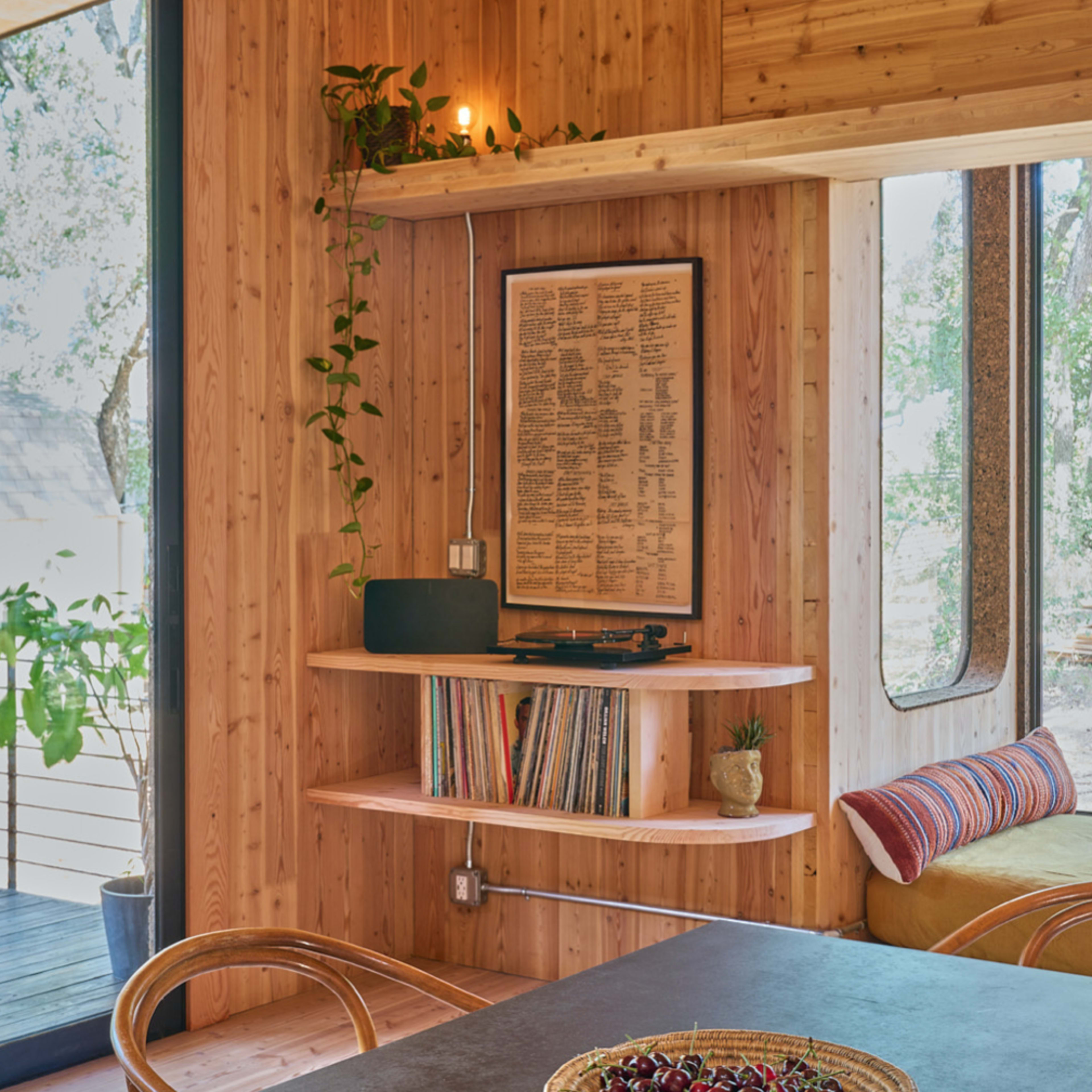 A wooden interior featuring shelves with books and plants, a record player, and a round bowl of cherries on a dark table.
