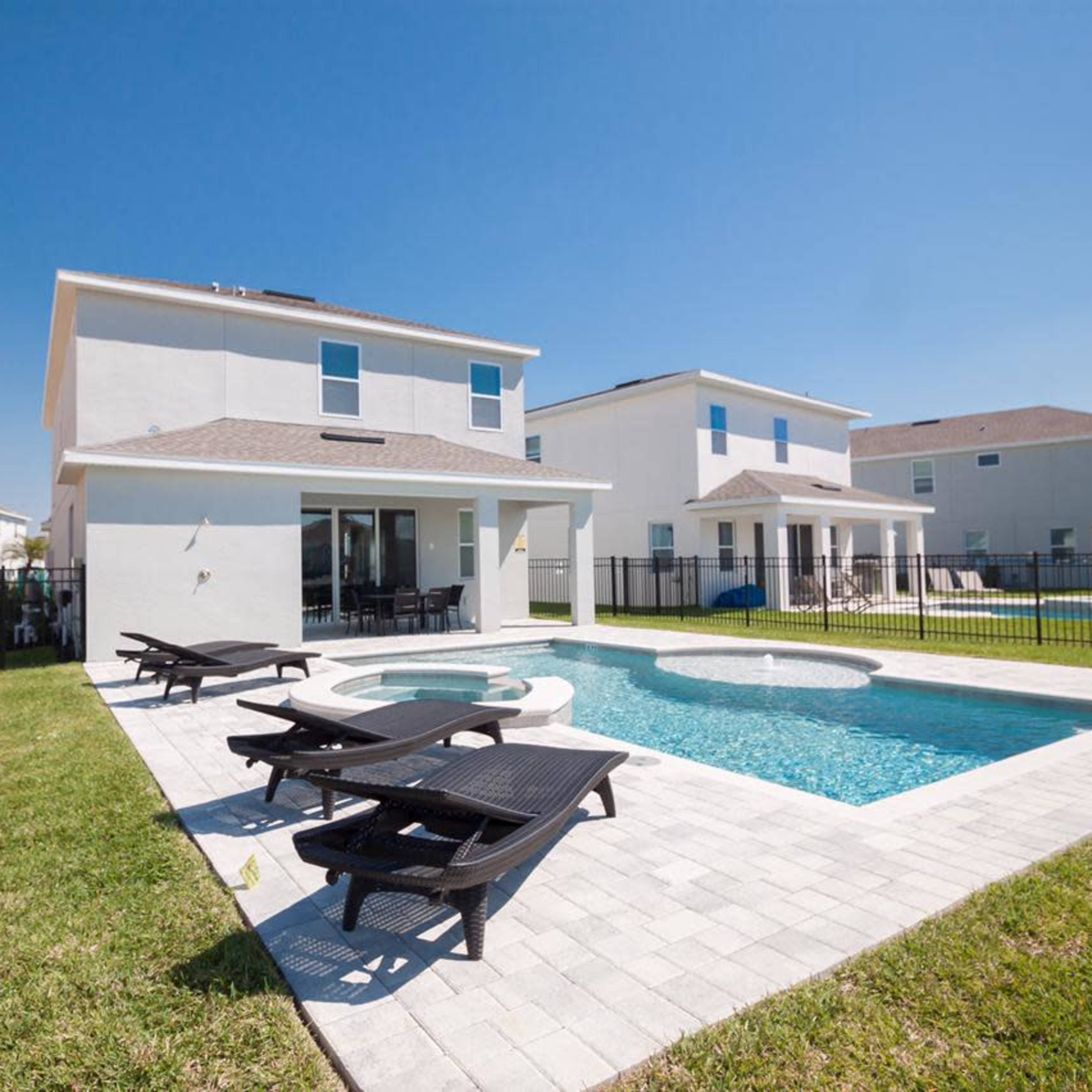 A backyard with a swimming pool surrounded by lounge chairs, two houses, and a fenced area under a clear blue sky.