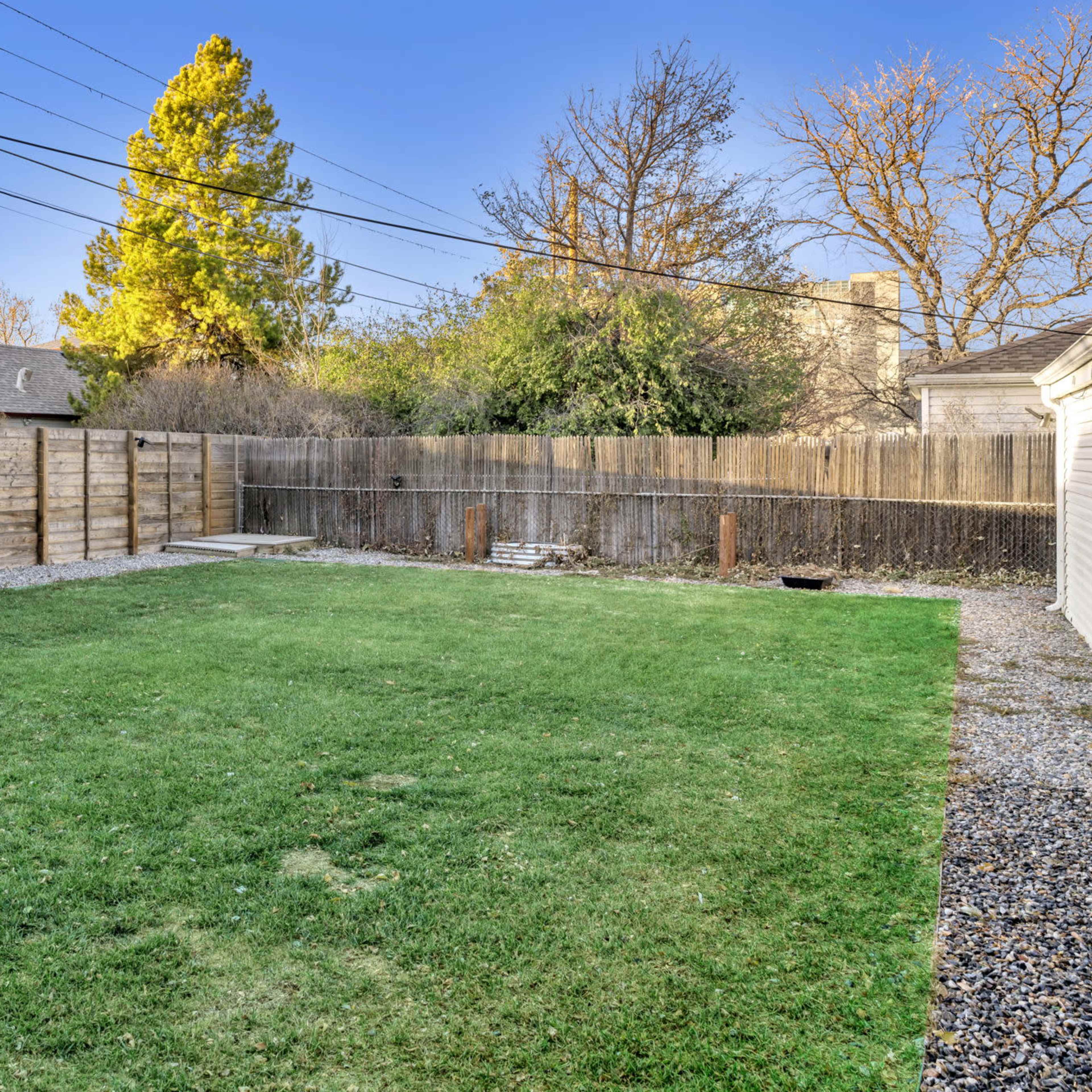 A grassy backyard enclosed by wooden fencing, with gravel borders and trees in the background.