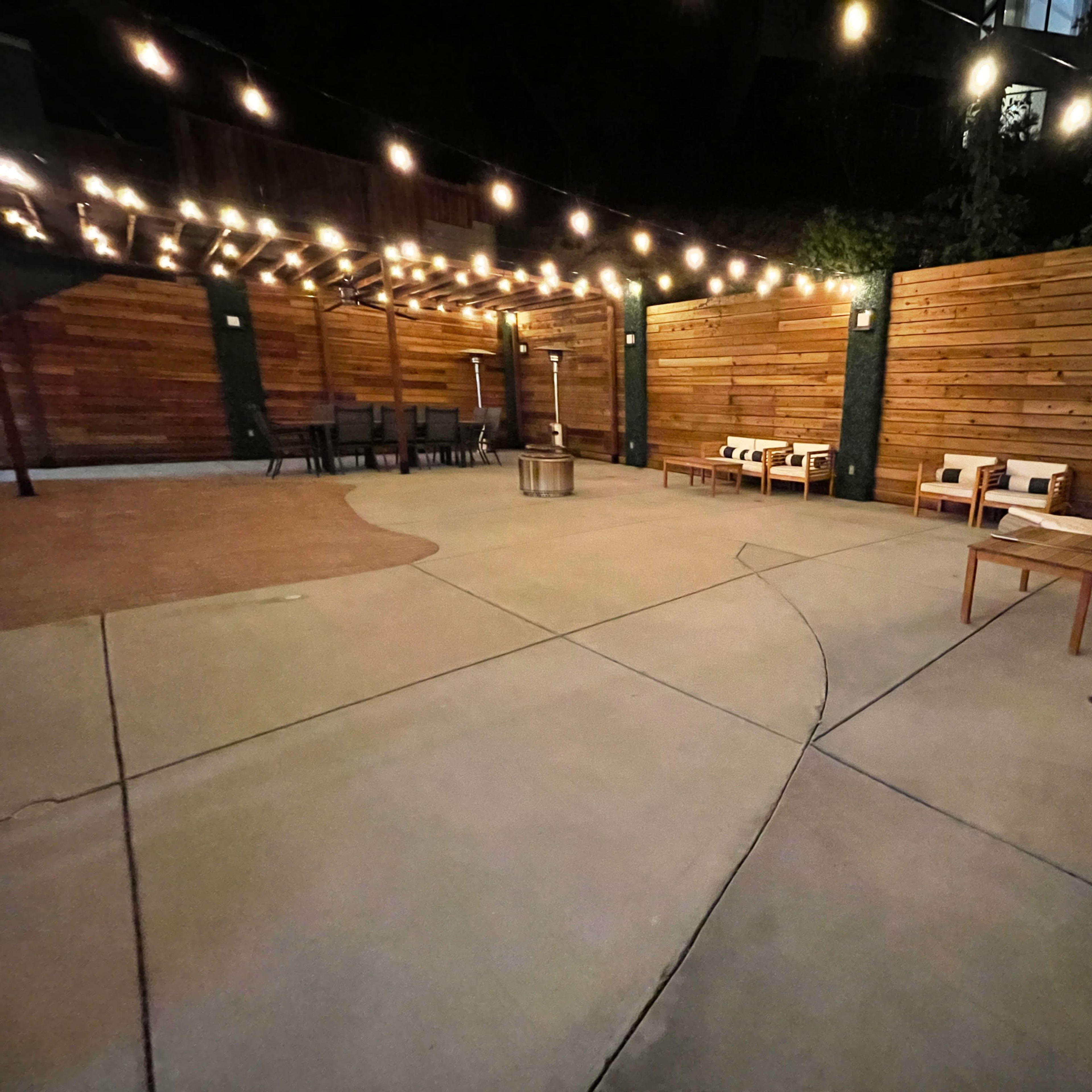 A dimly lit outdoor seating area with wooden walls, string lights overhead, and a few scattered chairs and tables on a concrete surface.