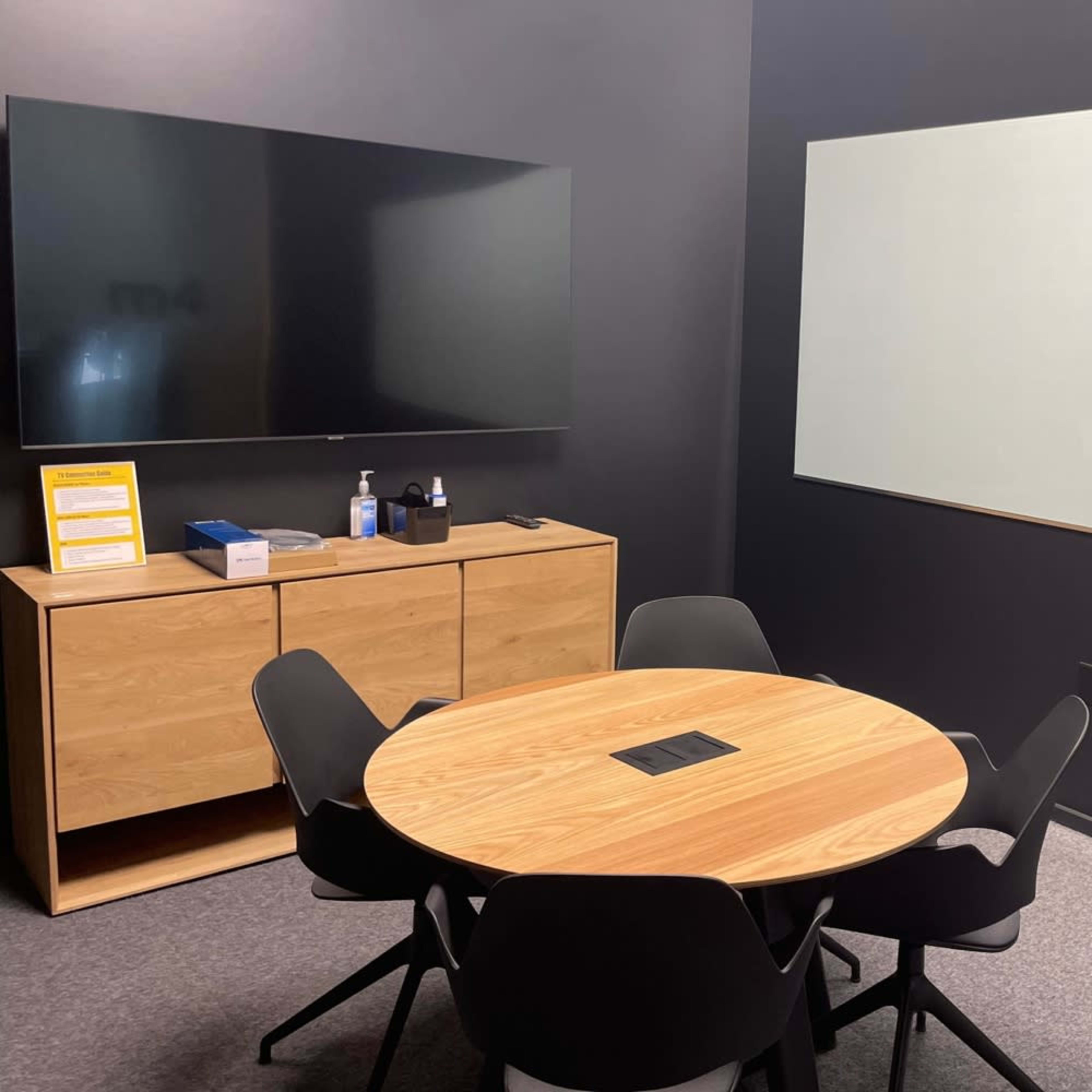 A meeting room features a round wooden table surrounded by black chairs, a wall-mounted flat-screen TV, and a blank whiteboard.