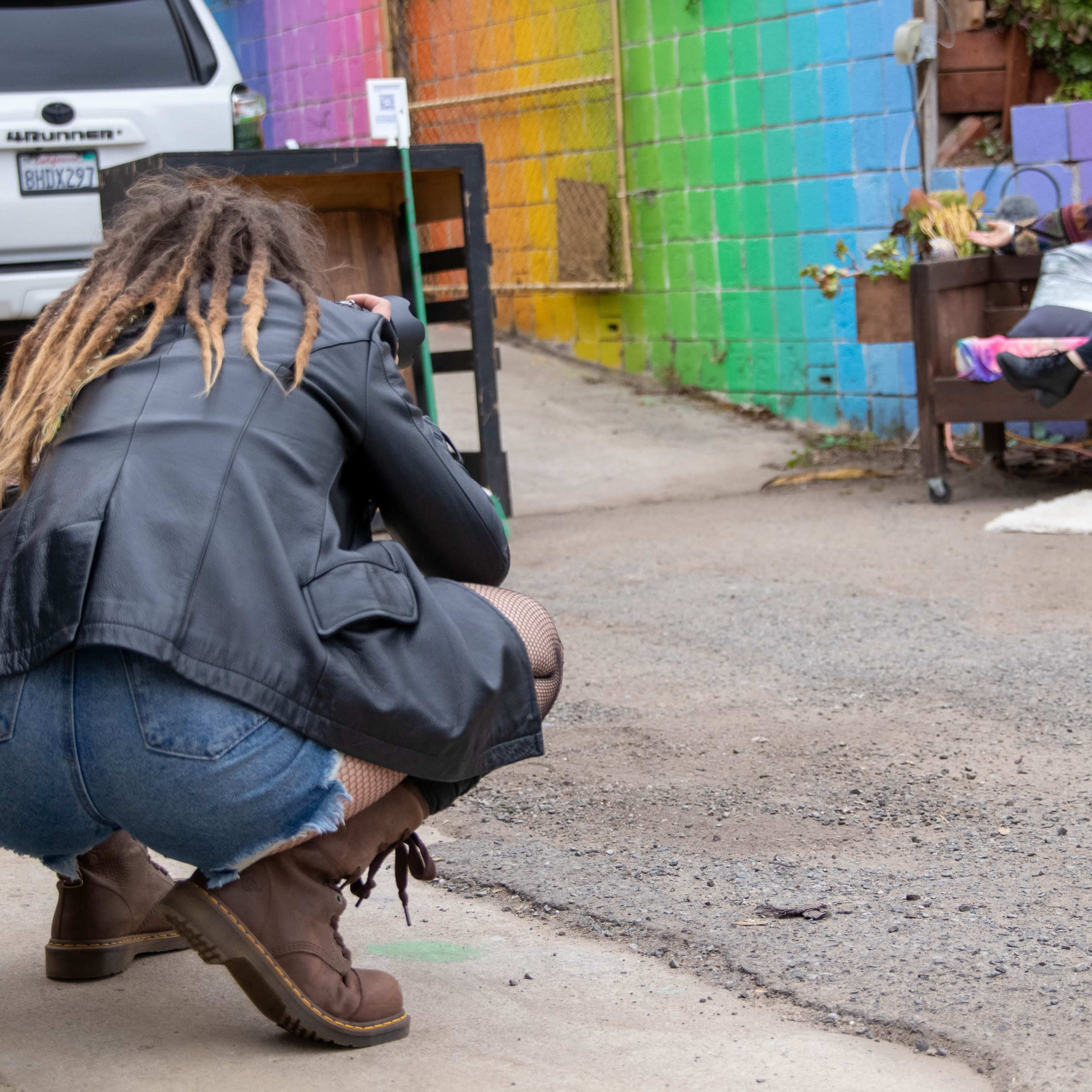 A photographer crouches on a street while capturing a model posing on a bench in front of a colorful mural.