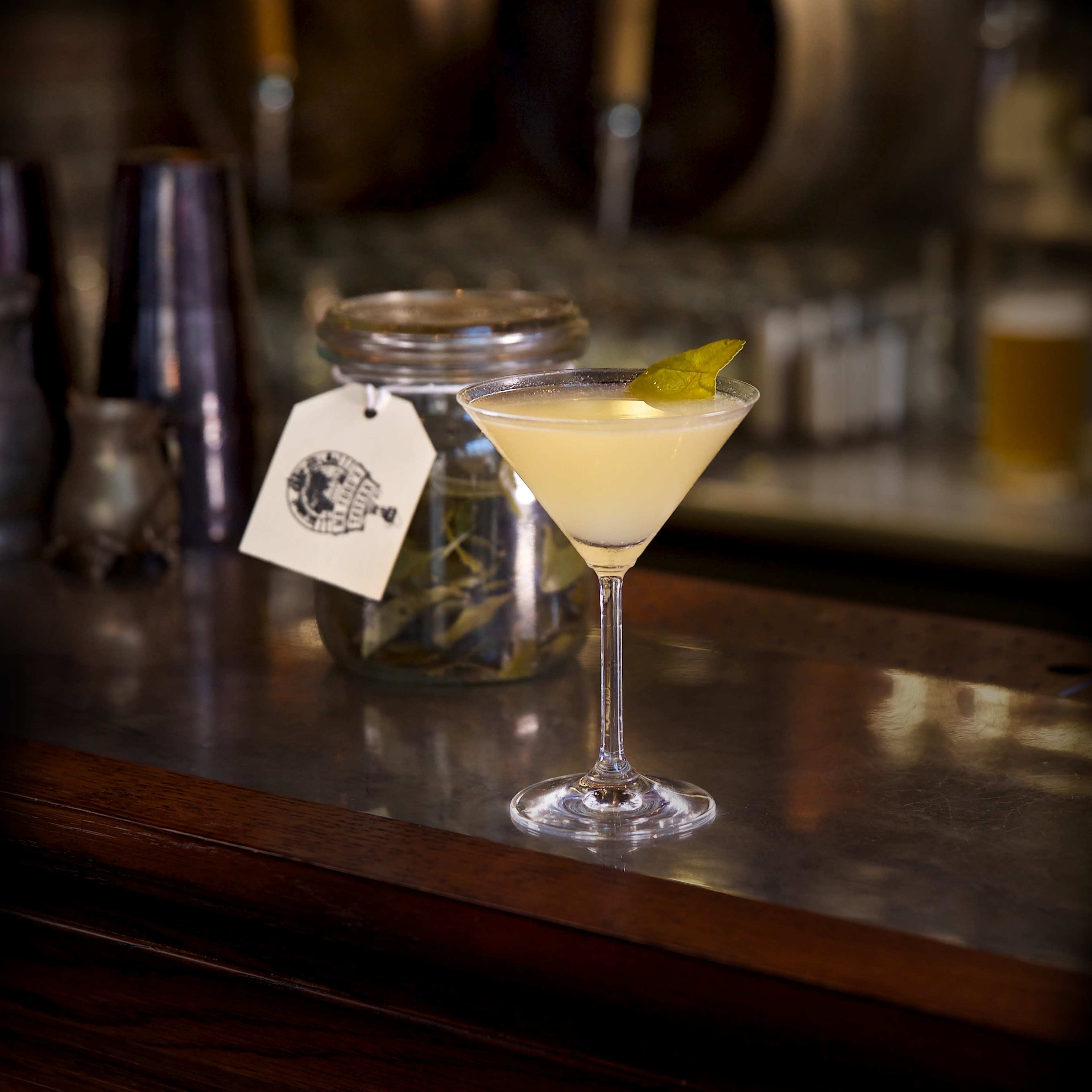 A martini with a slice of lemon is placed on a polished bar top next to a jar of herbs.