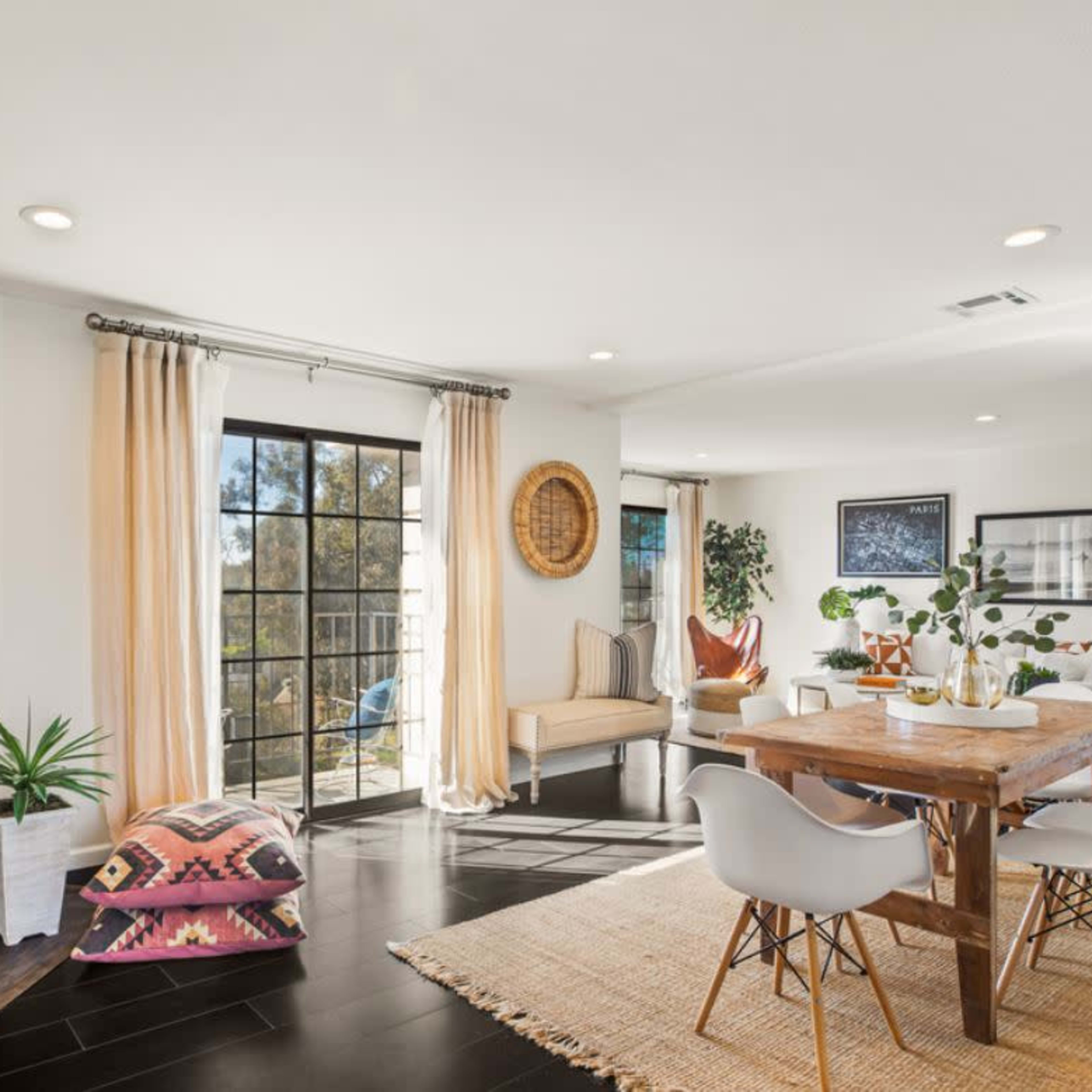 A light-filled living space featuring a dining area with a wooden table, white chairs, a fireplace, and large windows leading to an outdoor view.