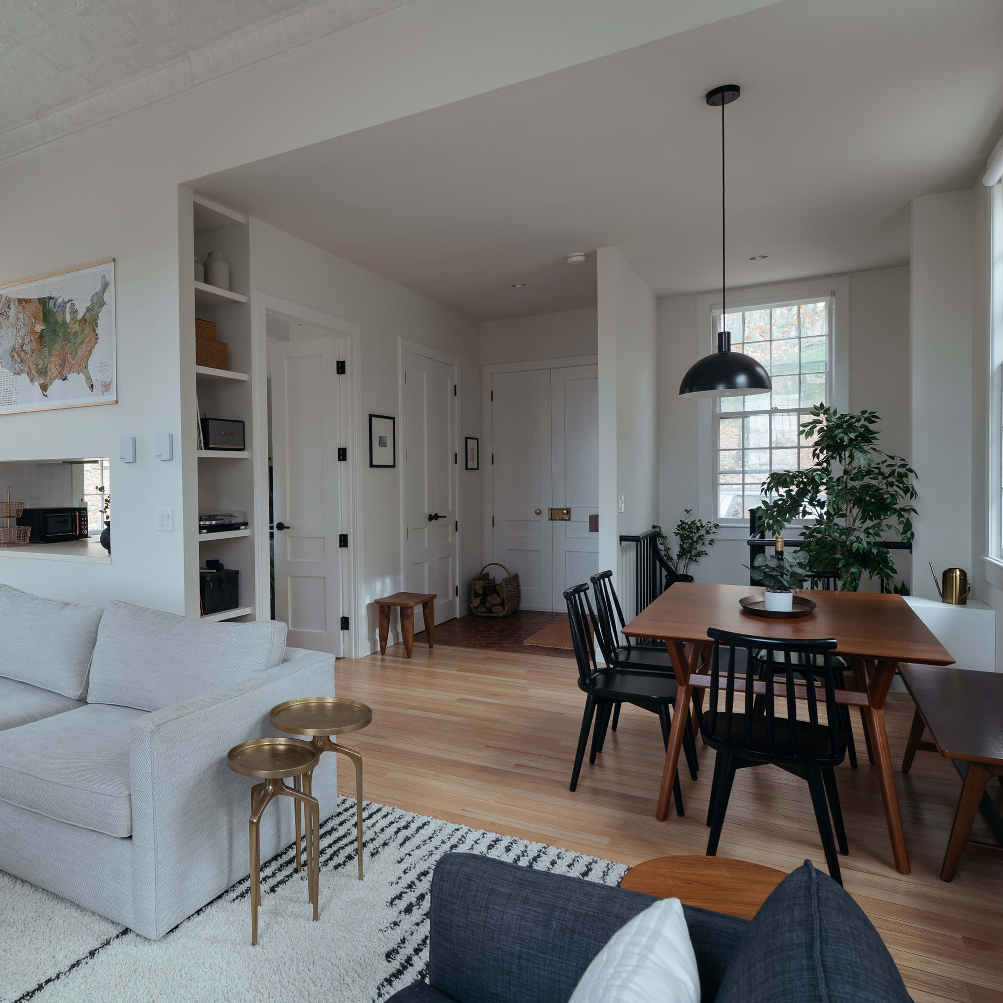 A modern living room with a light-colored sofa, a round coffee table, a dining area with a wooden table and chairs, and large windows allowing natural light to enter.