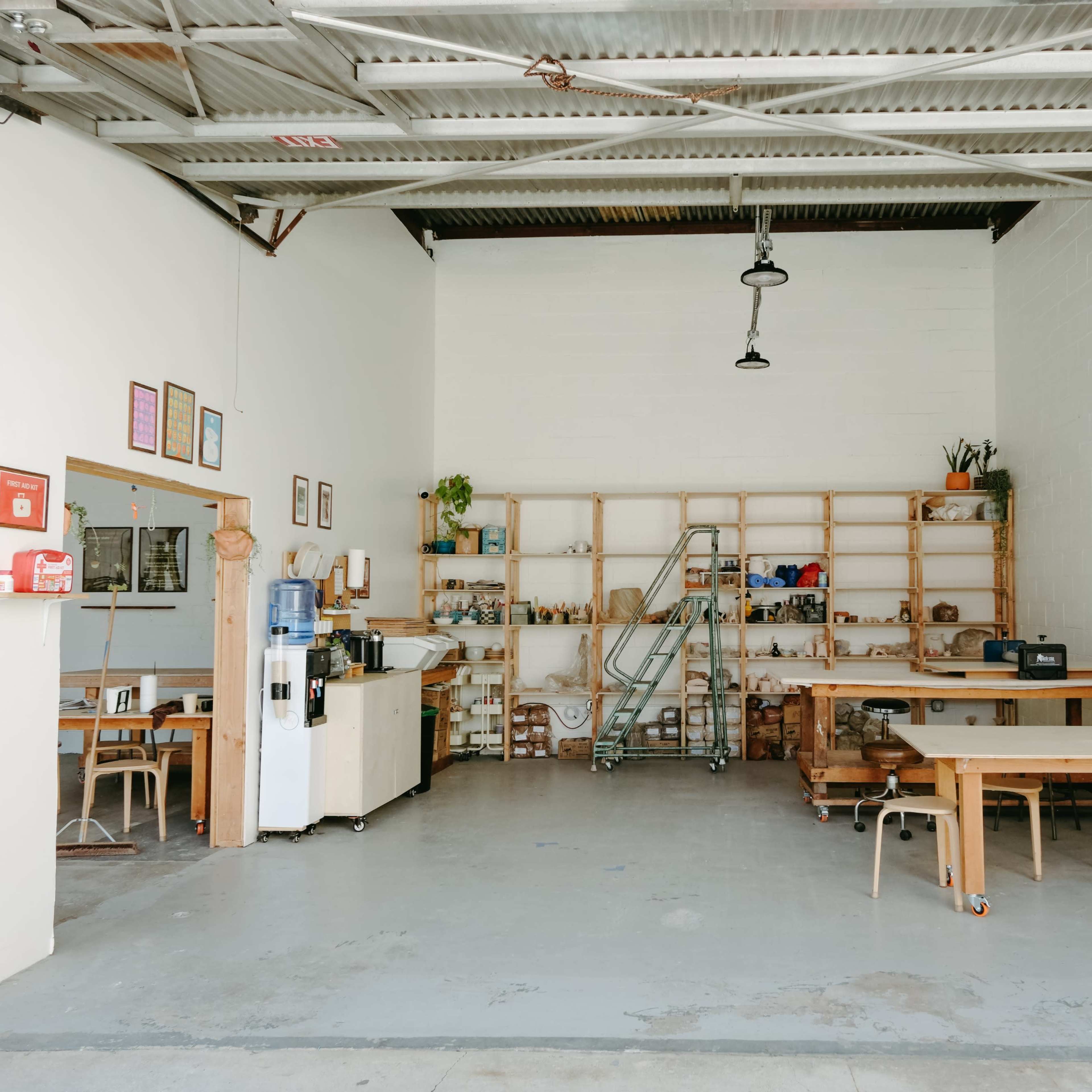 An empty, well-lit workspace featuring wooden shelves filled with items, a large table, chairs, and various appliances against a white wall.