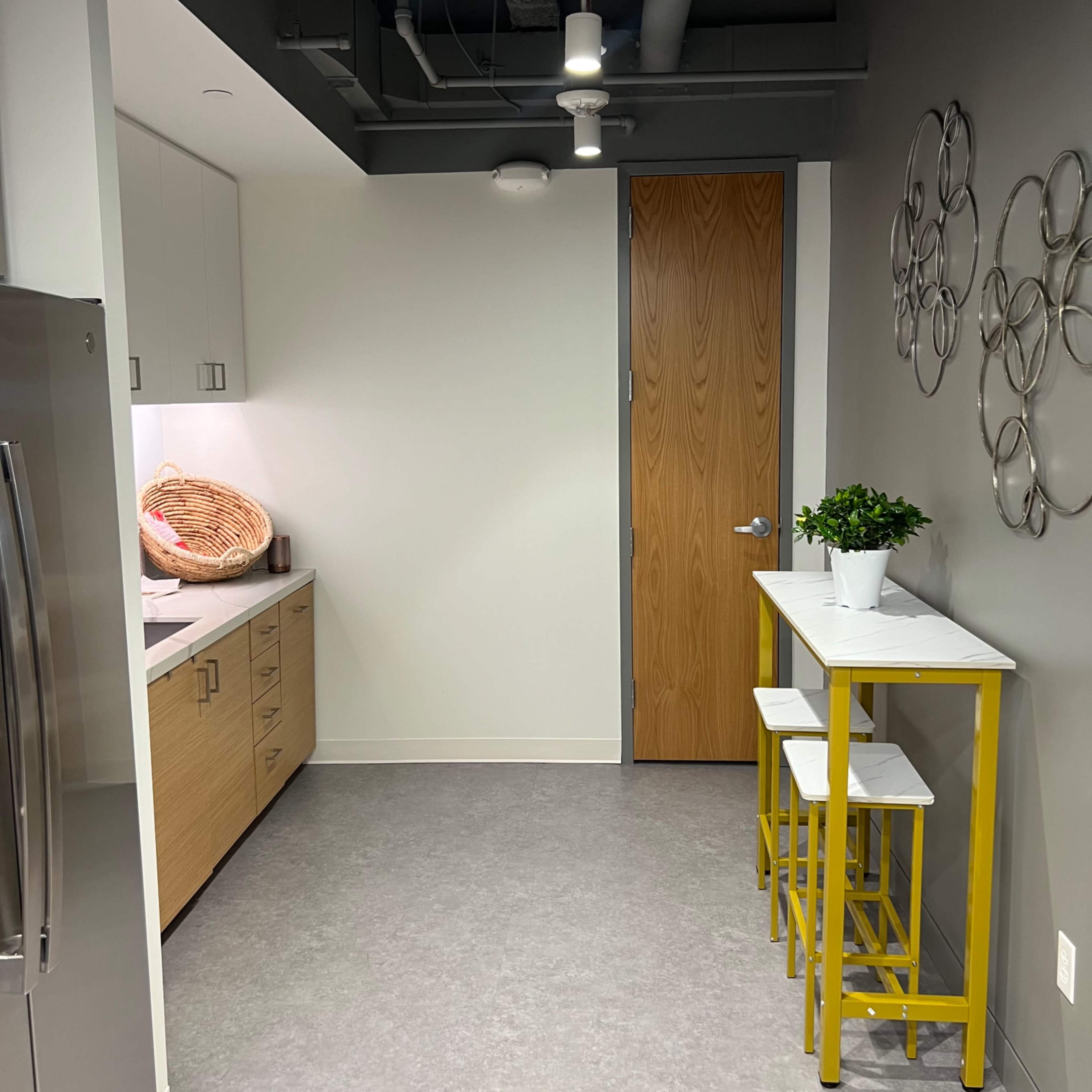 A modern kitchen area featuring a small dining table with yellow stools, a stainless steel refrigerator, and a door leading to another room.
