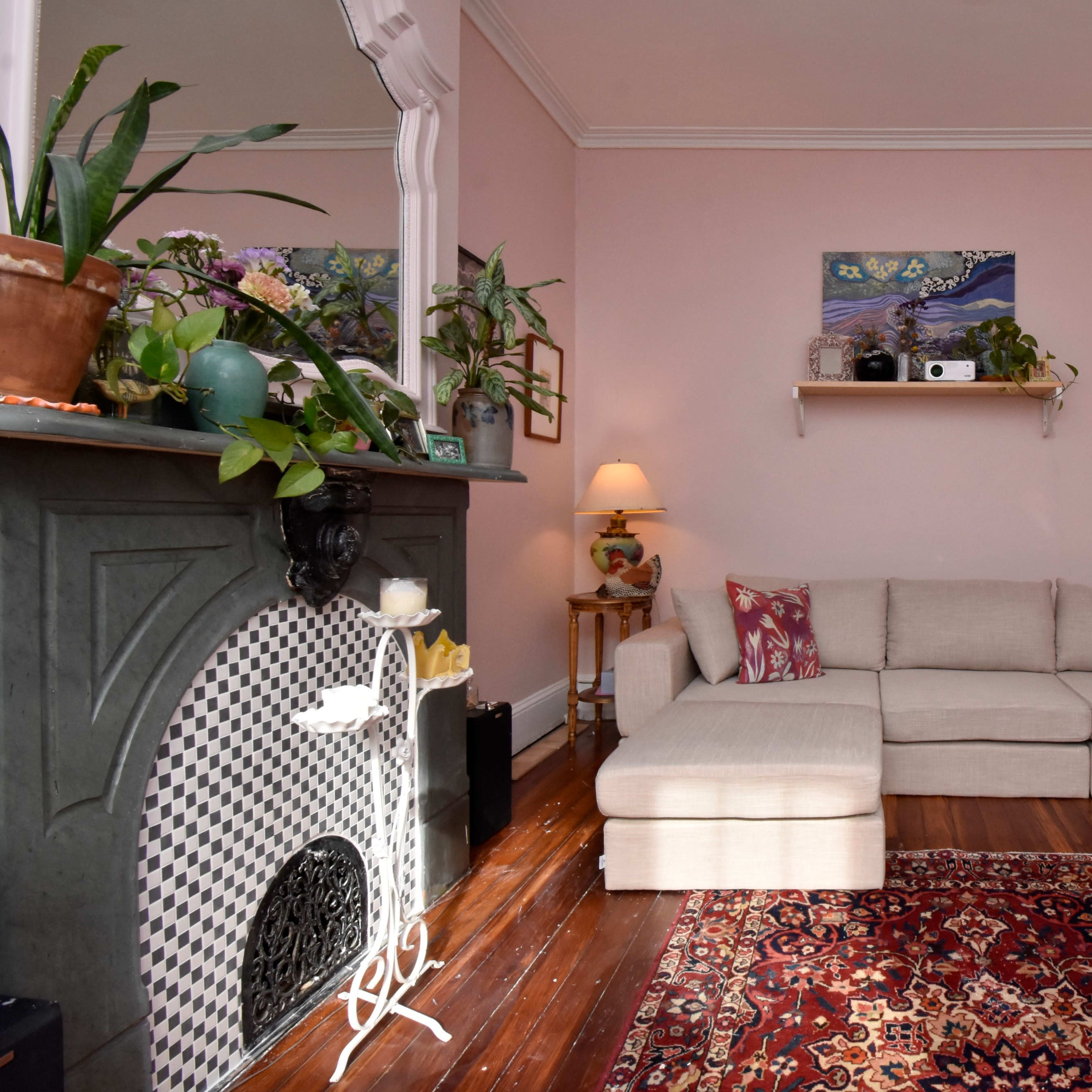 A living room with a light pink wall, a sectional sofa, a patterned rug, and decorative plants on shelves and furniture.