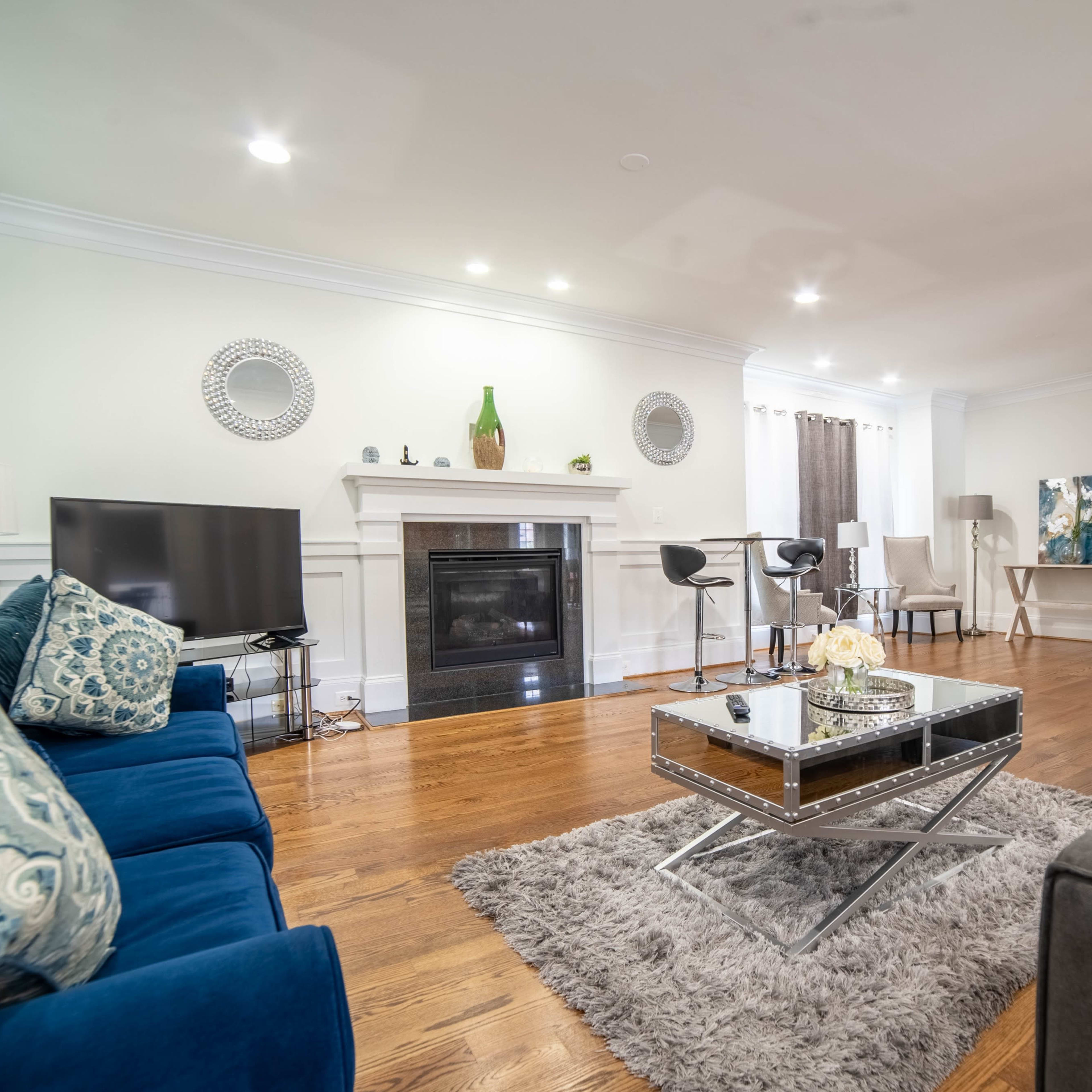 A modern living room features a fireplace, gray area rug, blue sofa, and a glass coffee table, with various seating arrangements and decorative items.