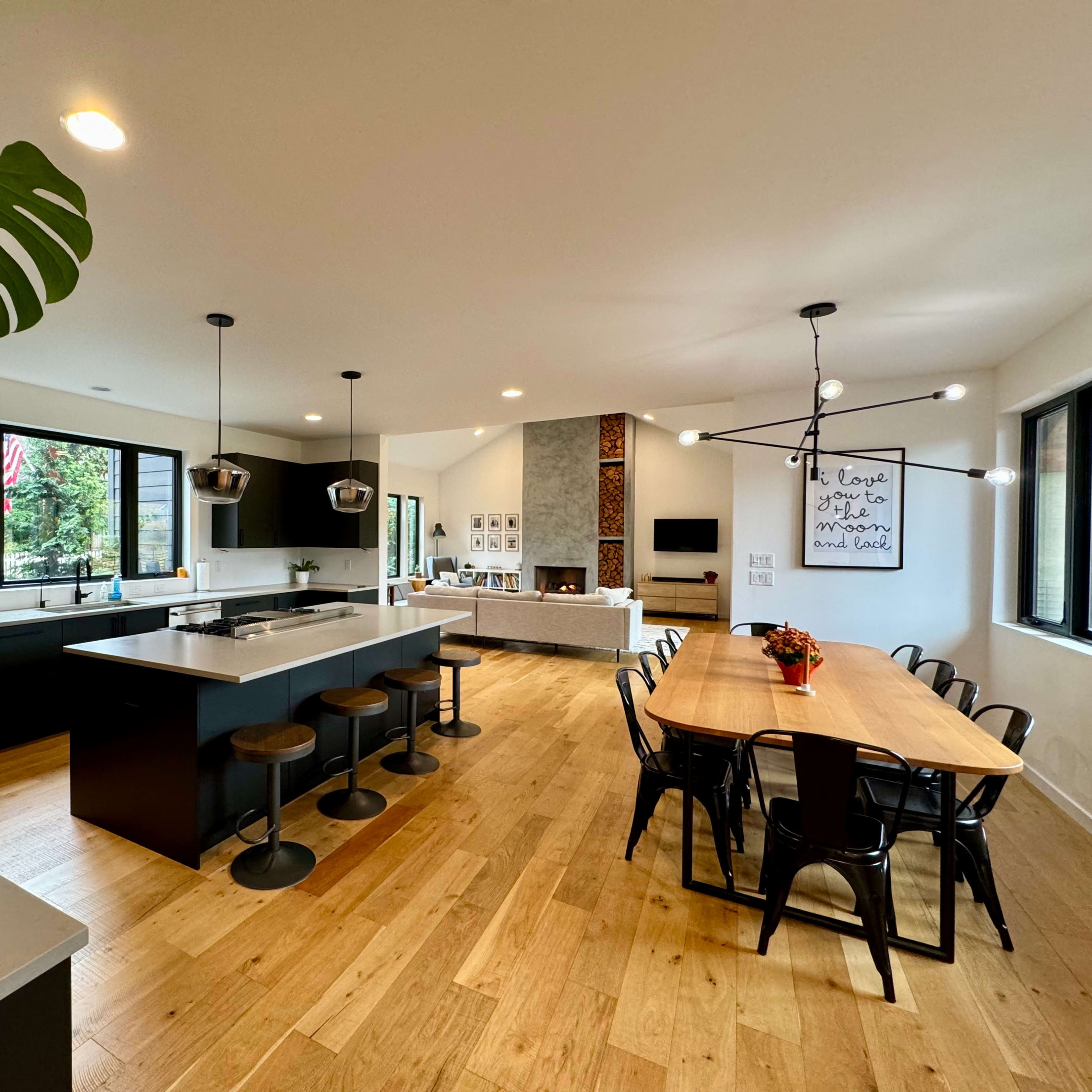 A modern kitchen and dining area featuring a central island with seating, dark cabinetry, a wooden dining table, and large windows allowing natural light.