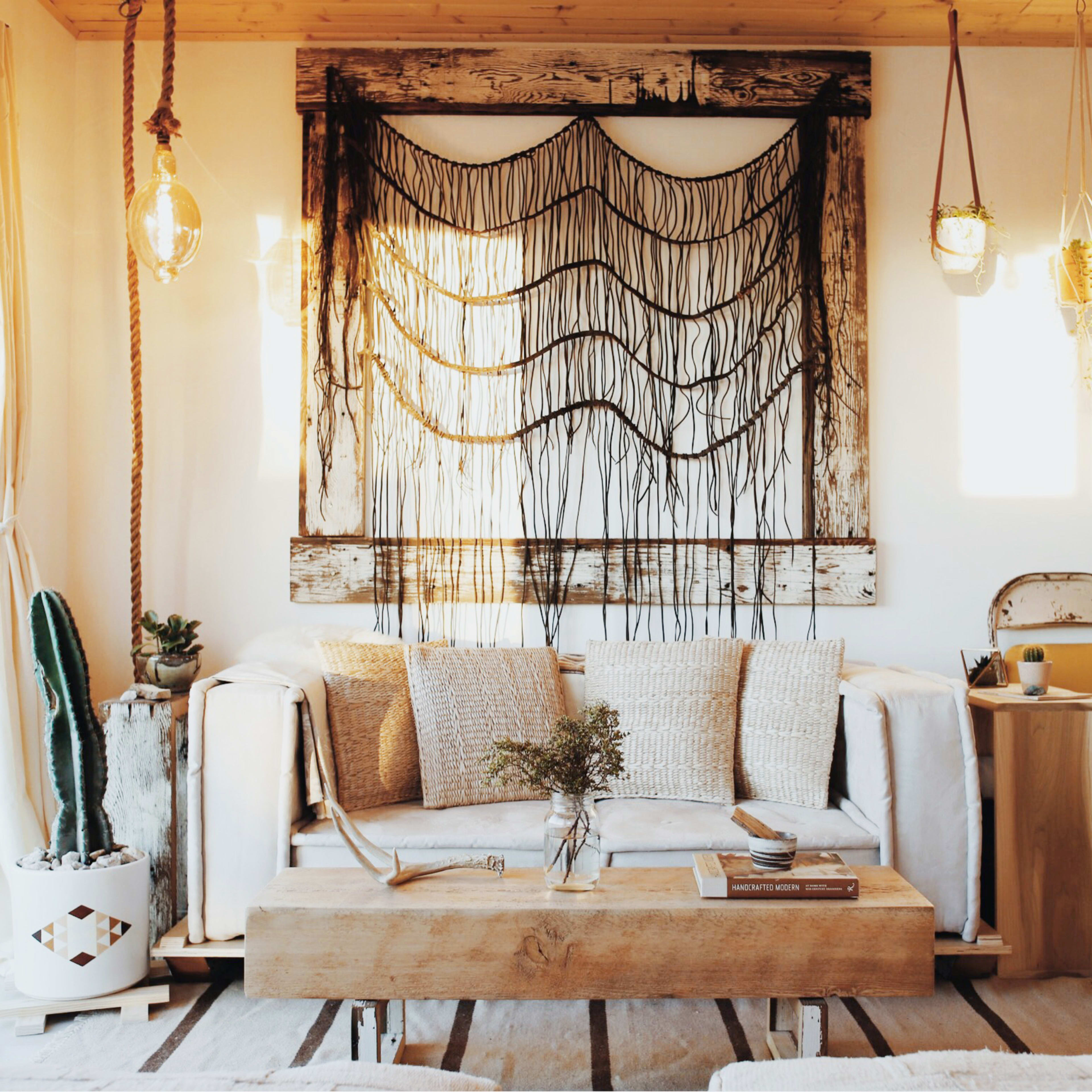 A living room with a white couch, a wooden coffee table, and a wall adorned with a macramé art piece, illuminated by natural light from large windows.