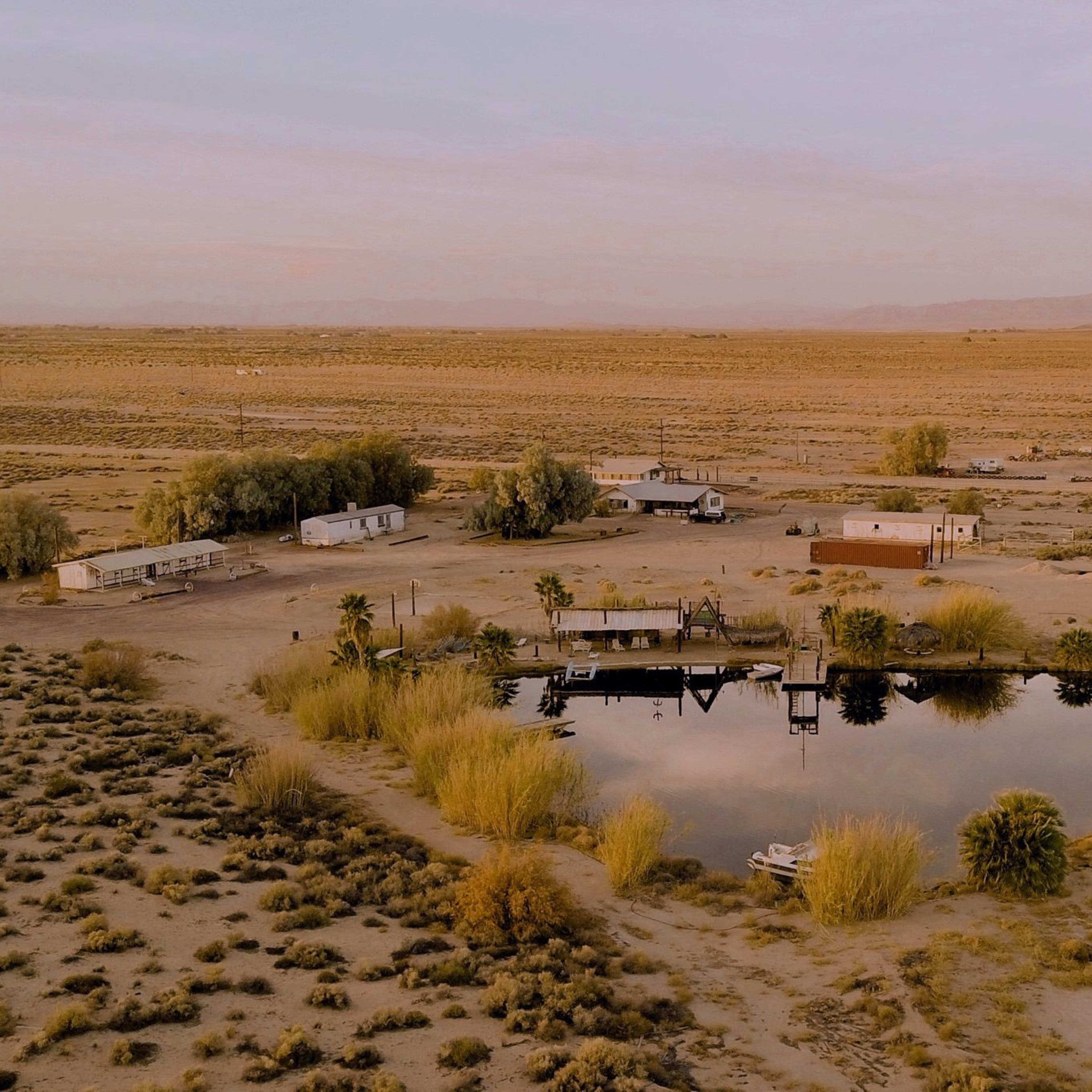 A remote desert landscape with several trailers and a pond surrounded by sparse vegetation.