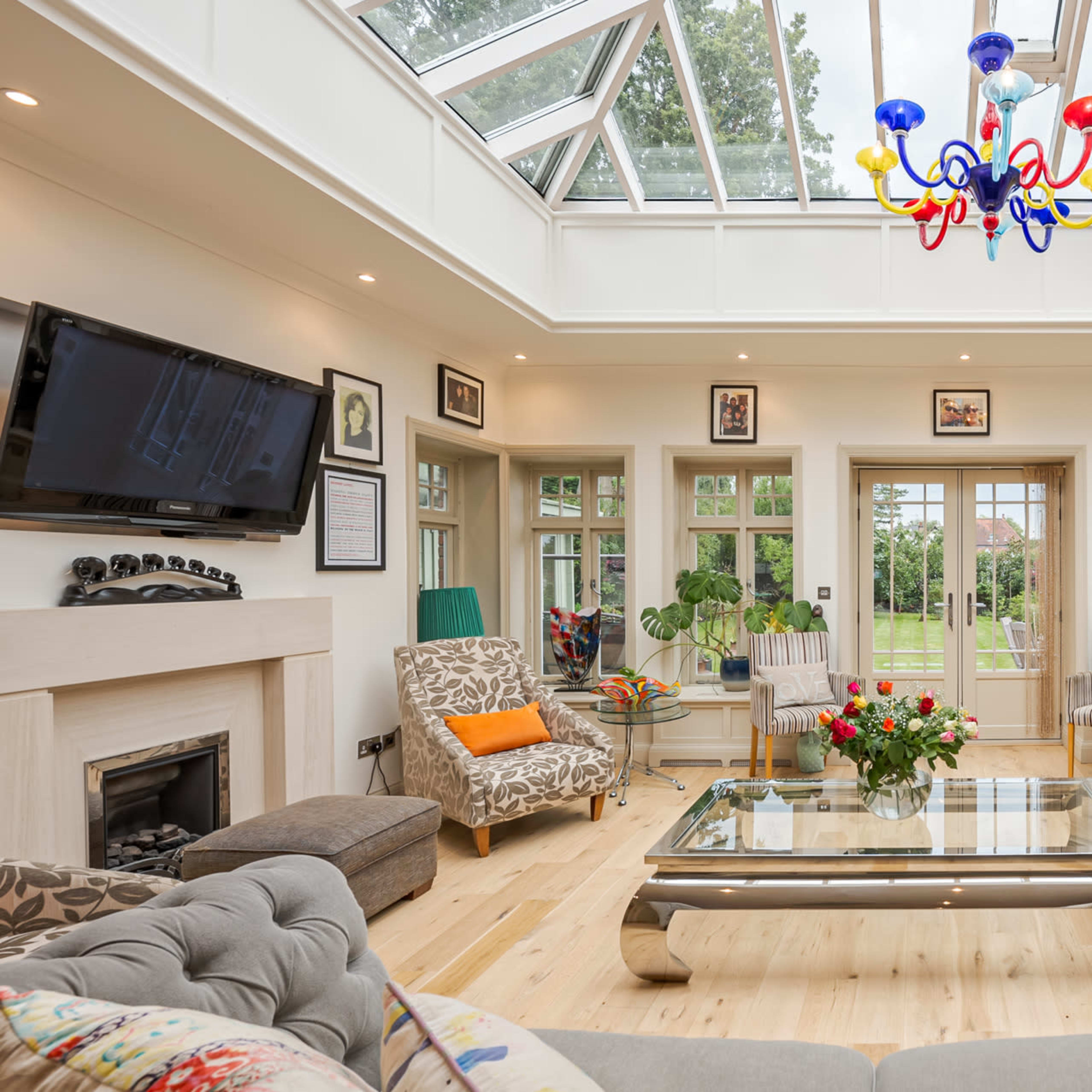 A bright conservatory with a glass ceiling, featuring a spacious seating area with a coffee table and a view of a garden outside.