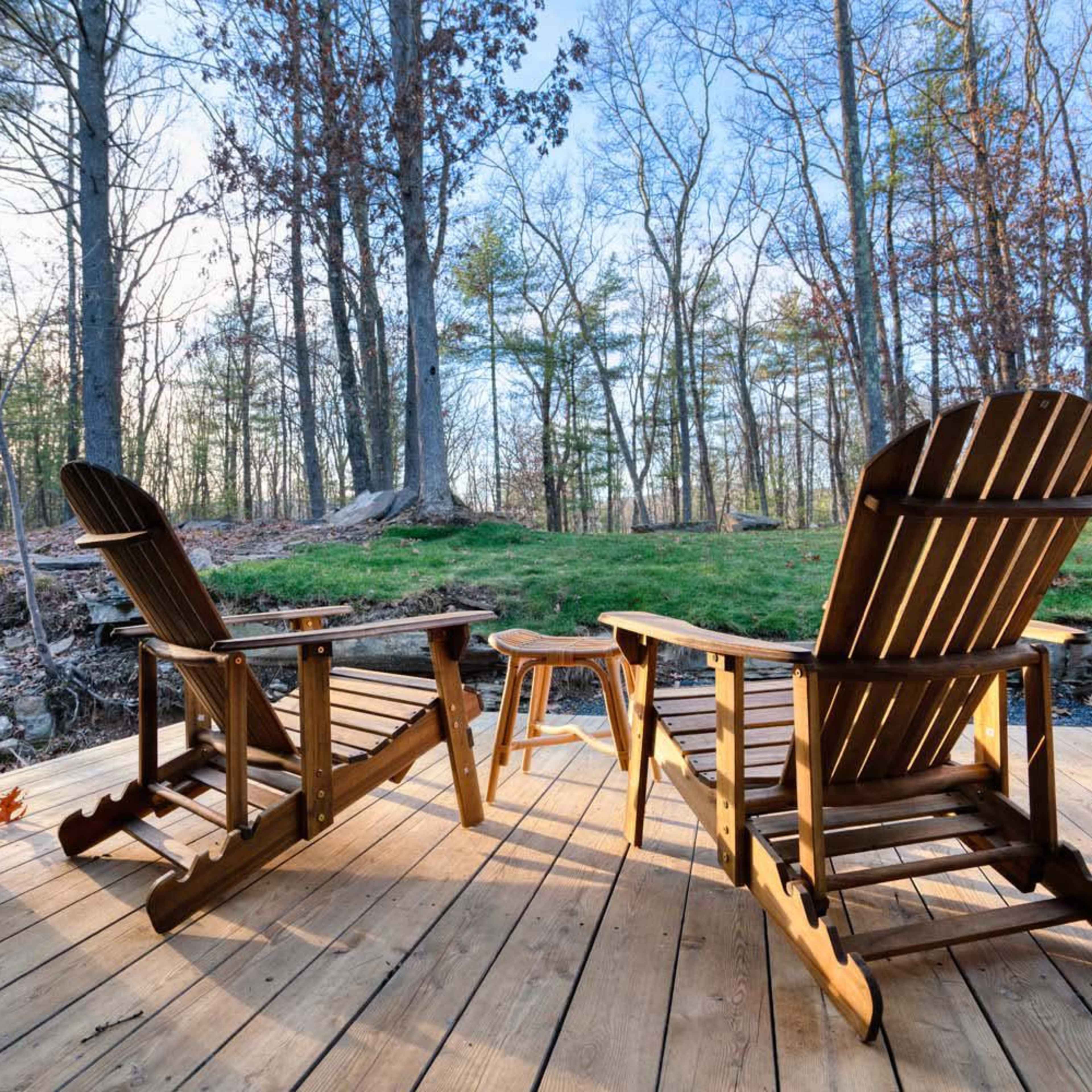 Two wooden Adirondack chairs and a small table are positioned on a wooden deck overlooking a grassy area surrounded by trees.