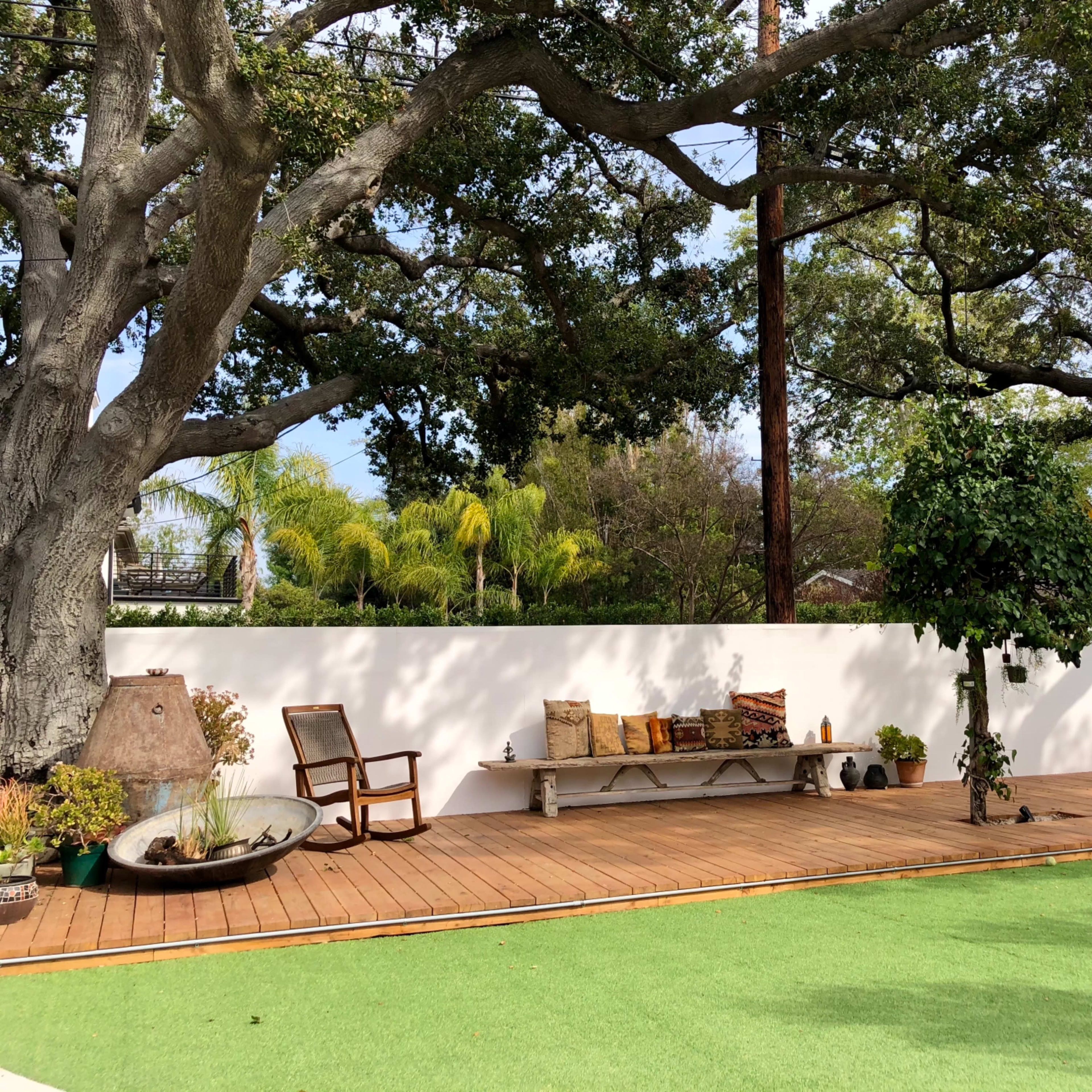 A wooden deck with a bench, a small red table and chairs, and a green lawn bordered by trees and a white wall.
