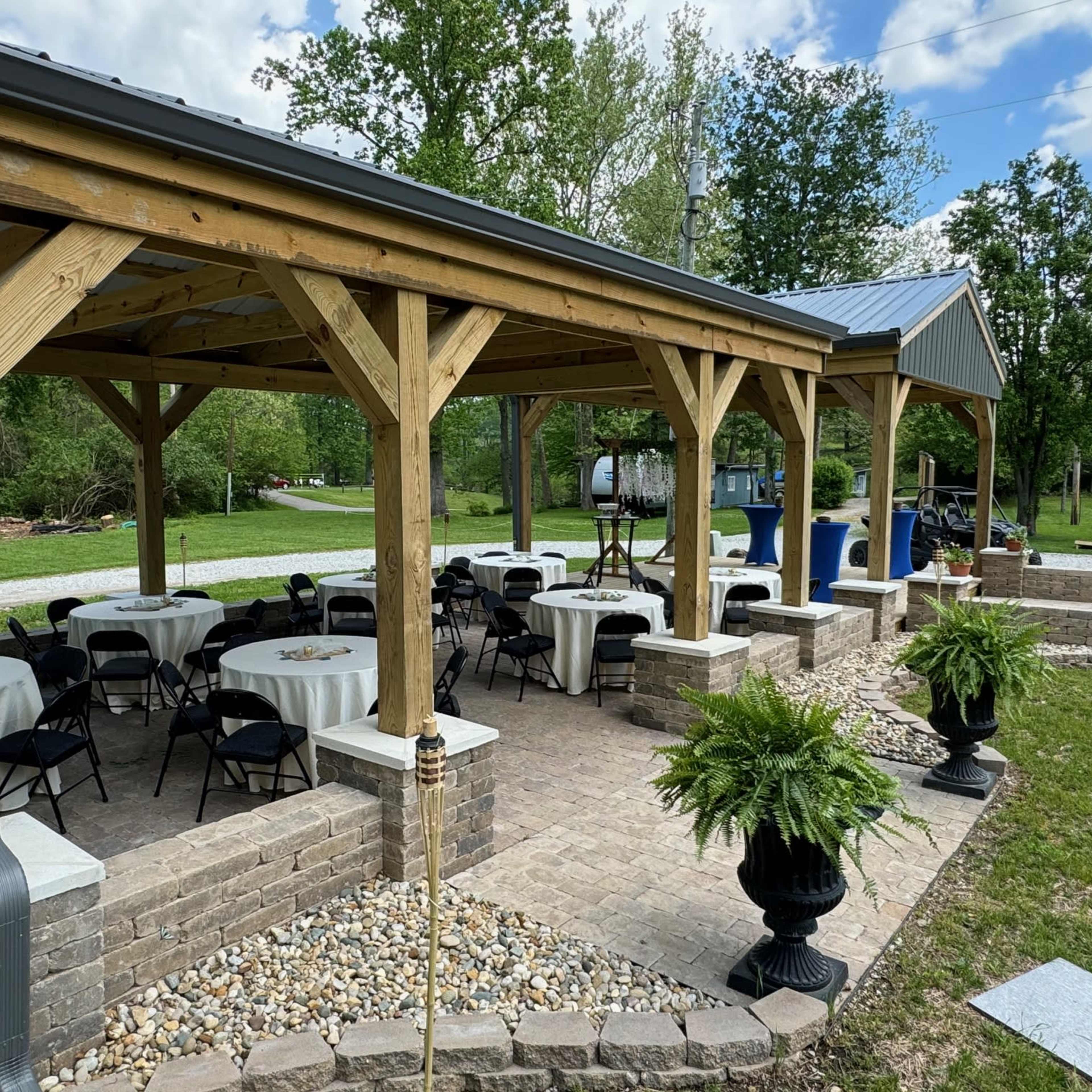 A spacious outdoor pavilion with tables and chairs is set up on a stone patio, surrounded by greenery.
