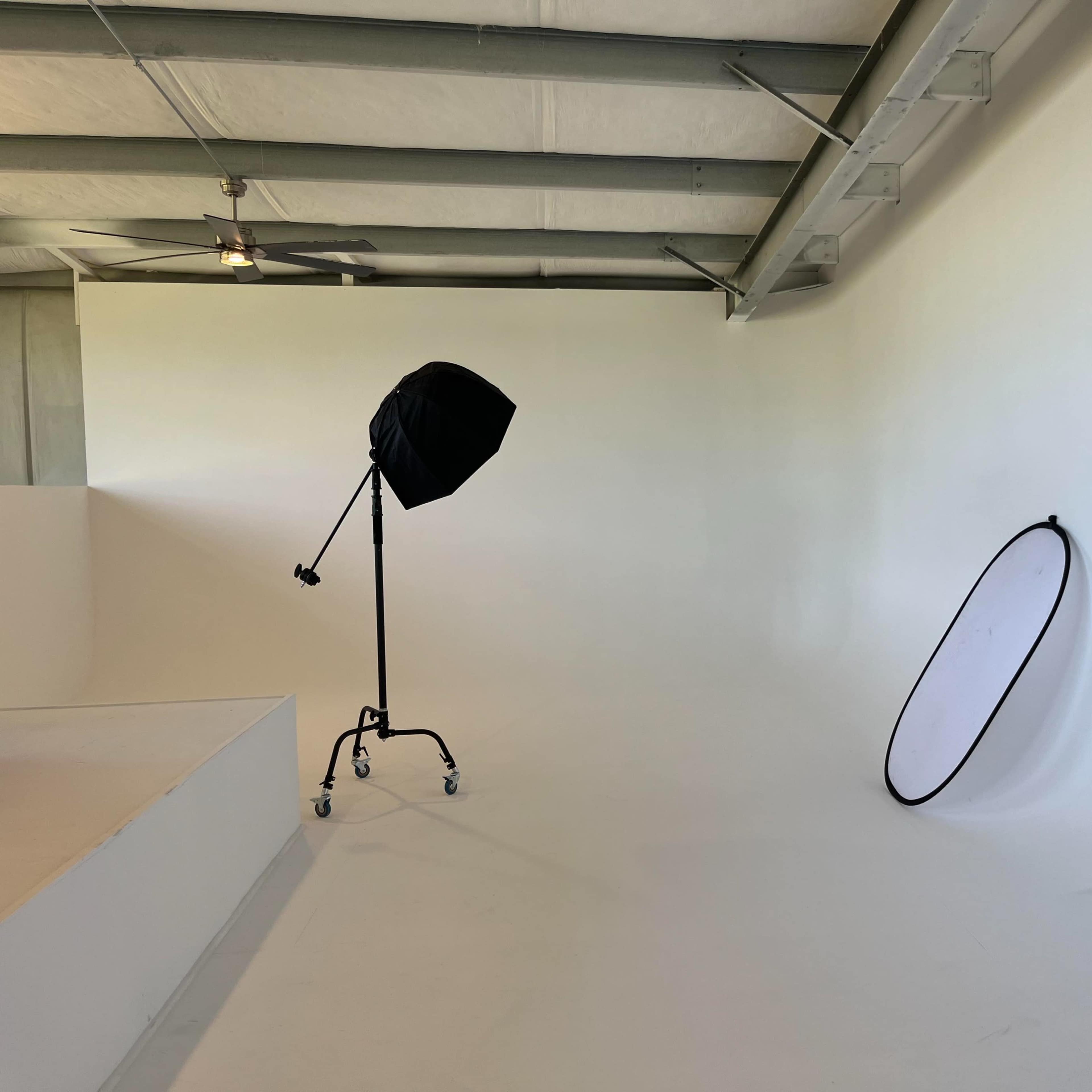 The scene shows a minimalistic photo studio with a softbox light and a large reflector leaning against a white wall.
