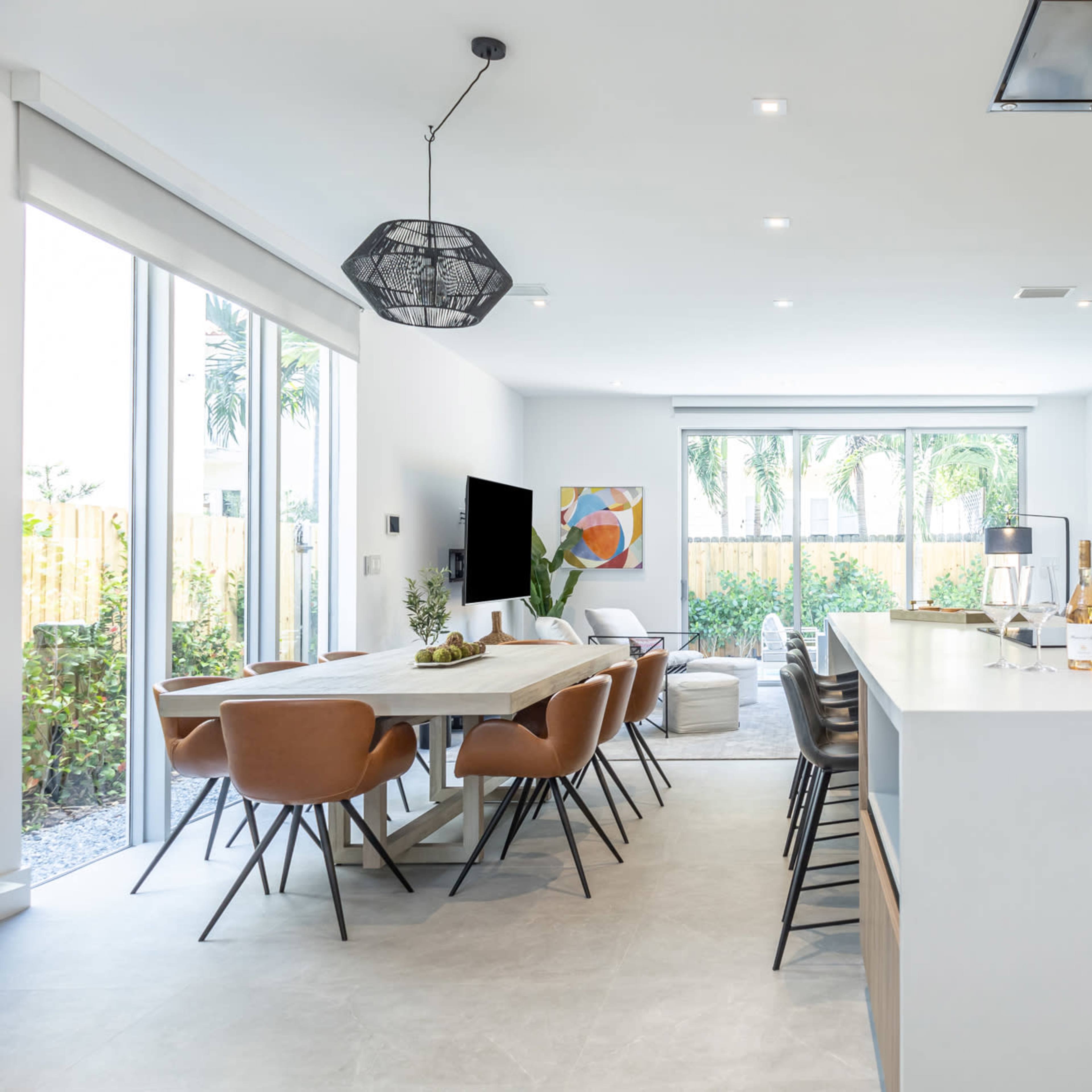 A modern kitchen and dining area features a large table, pendant lighting, and large glass doors opening to an outdoor space with greenery.