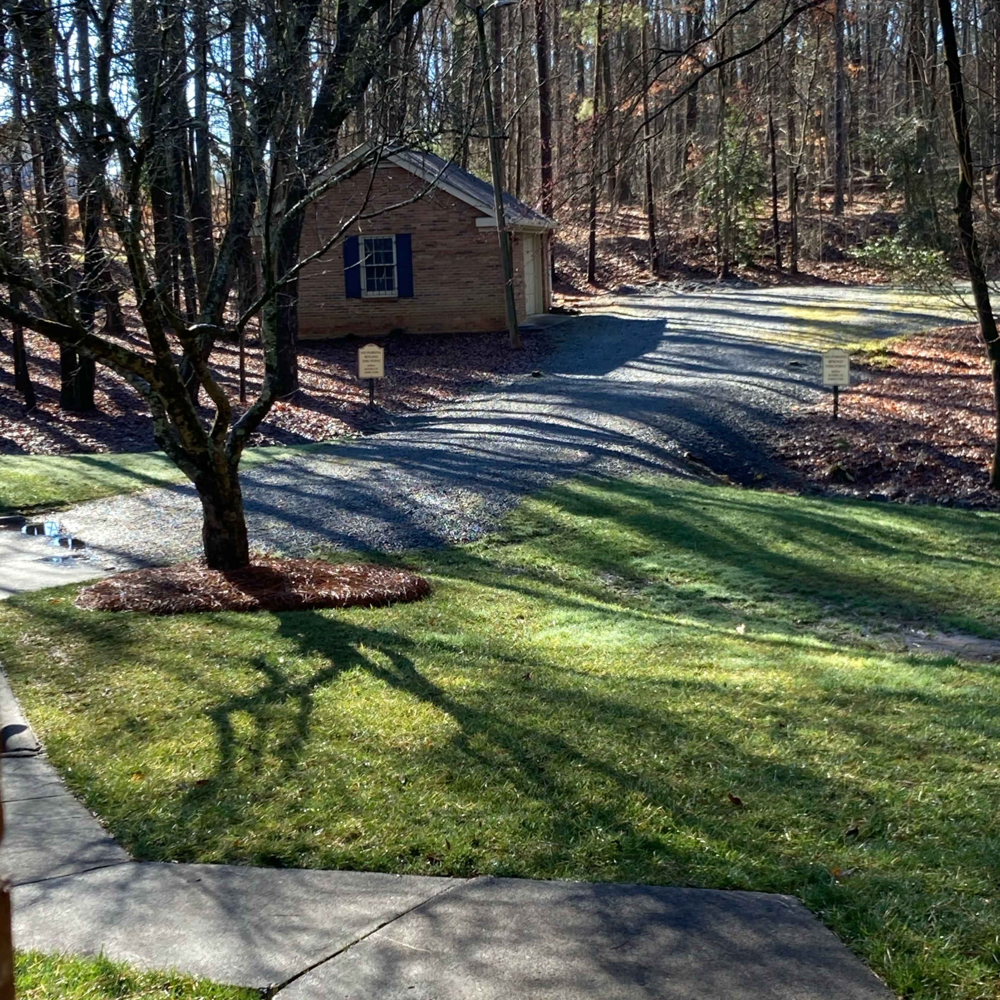 A small brick shed is situated at the end of a gravel driveway, surrounded by trees and a manicured lawn.