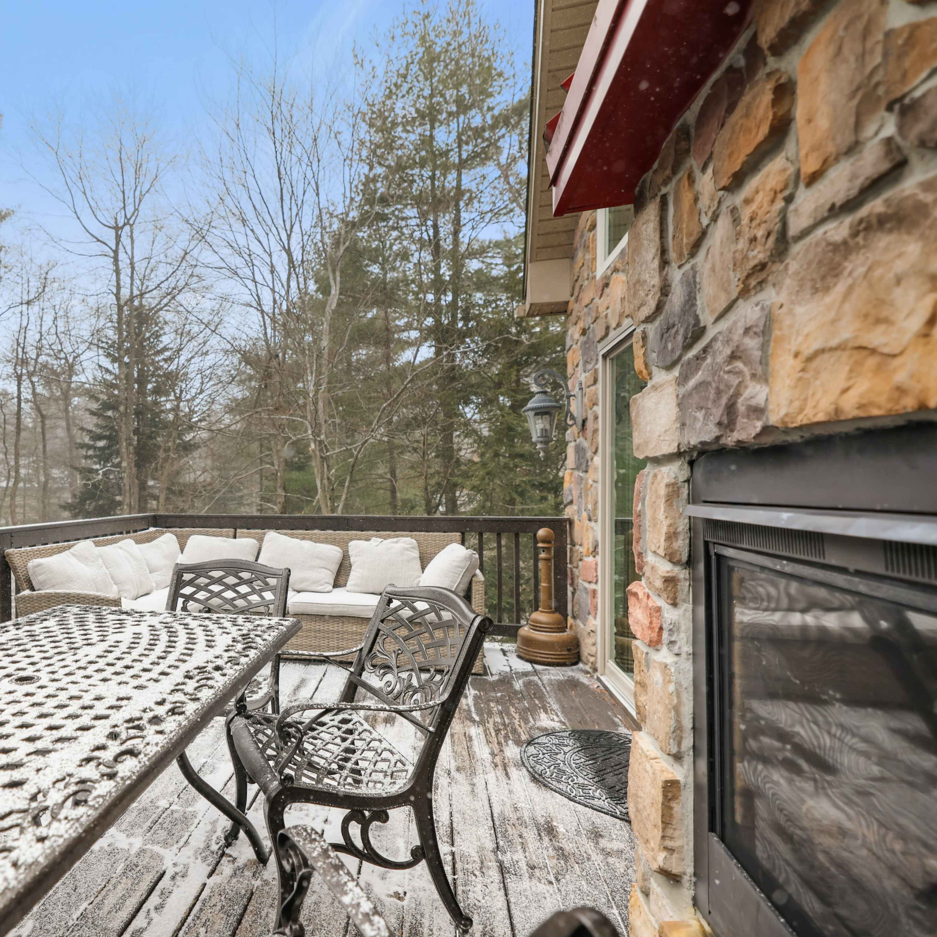 A dining table with metal chairs is set on a snow-covered deck adjacent to a stone wall and surrounded by trees in a winter landscape.
