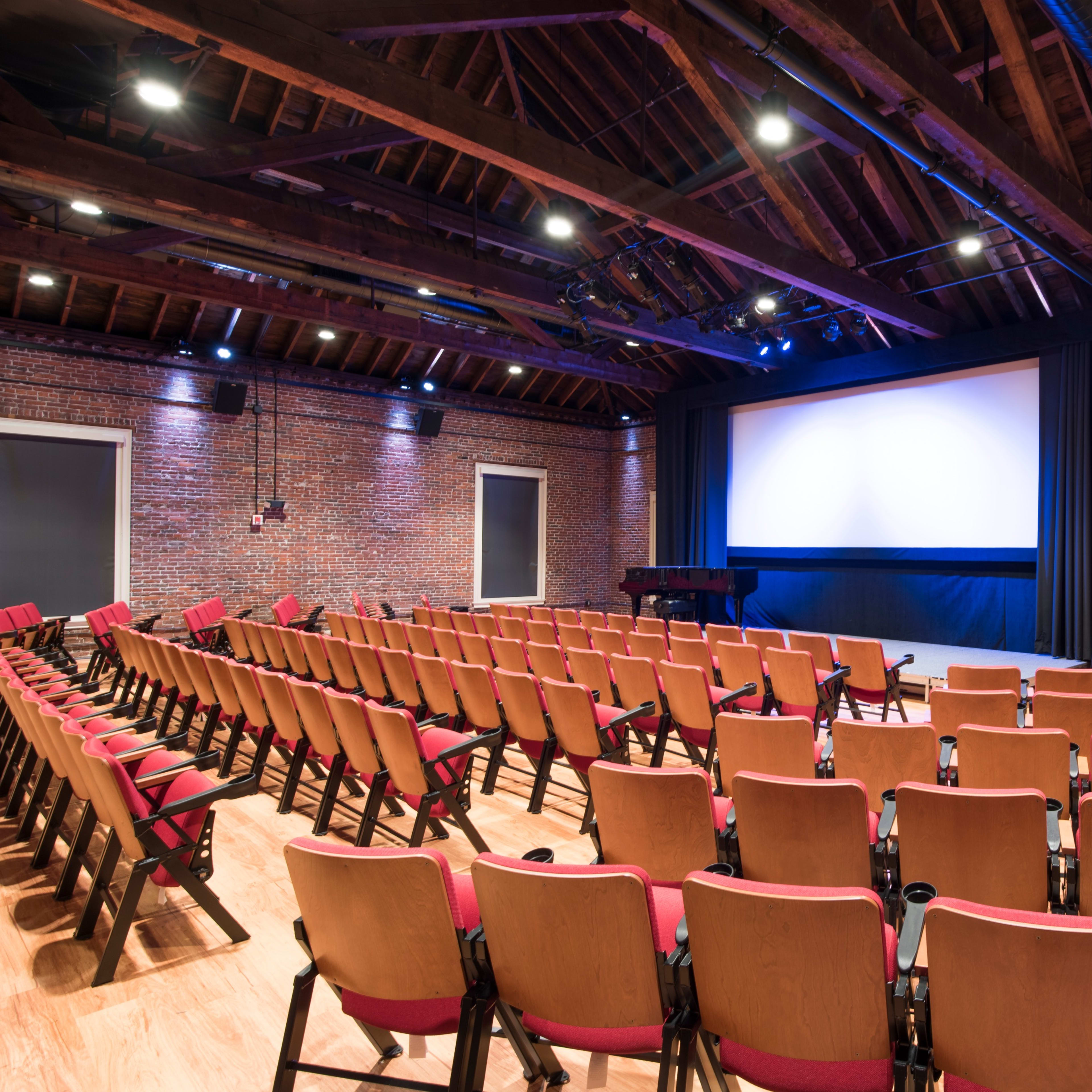 A small theater features rows of red cushioned seating facing a large projection screen, set against a backdrop of brick walls and wooden beams.
