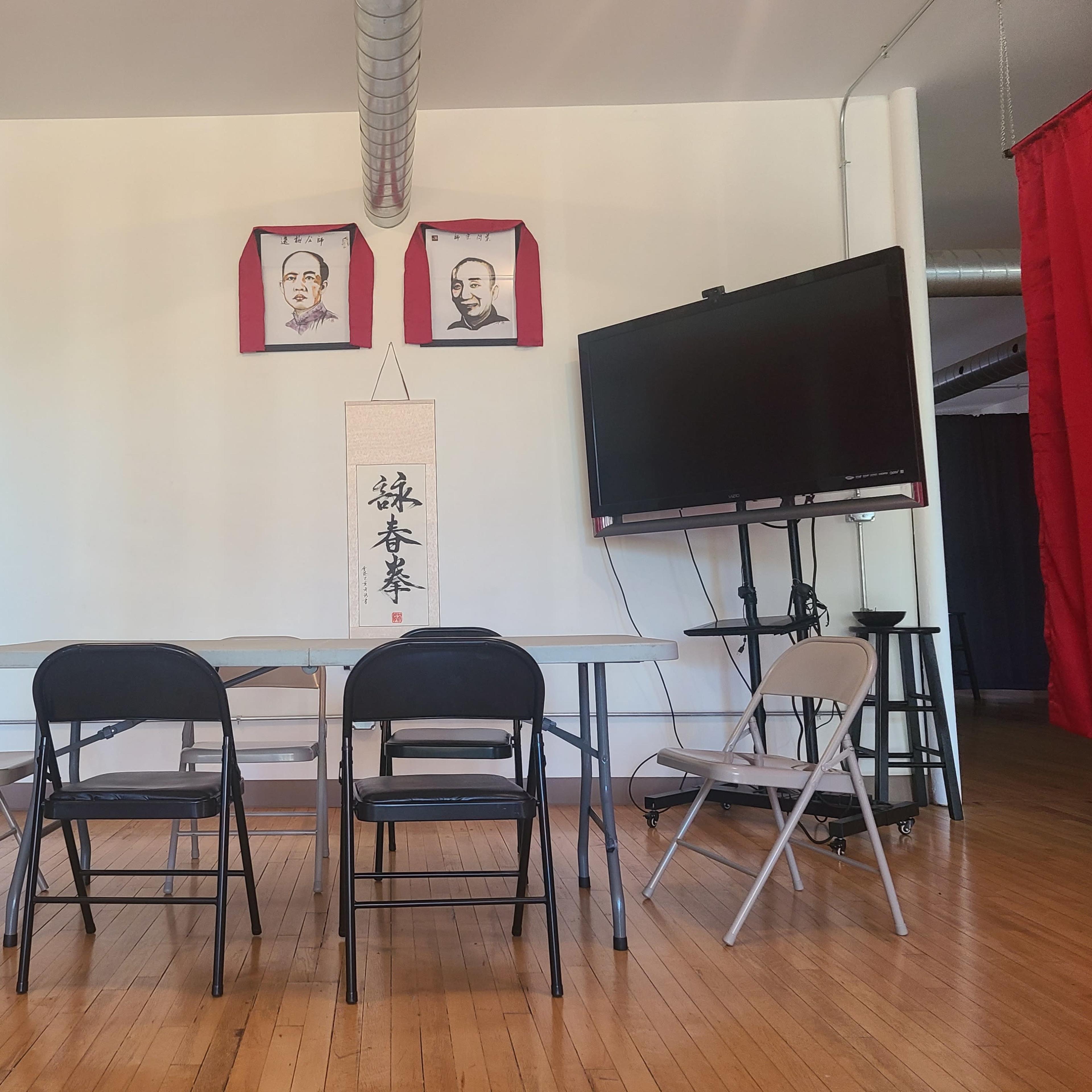 A meeting room features a rectangular table surrounded by black and white folding chairs, with two portraits on the wall and a large screen on the right.